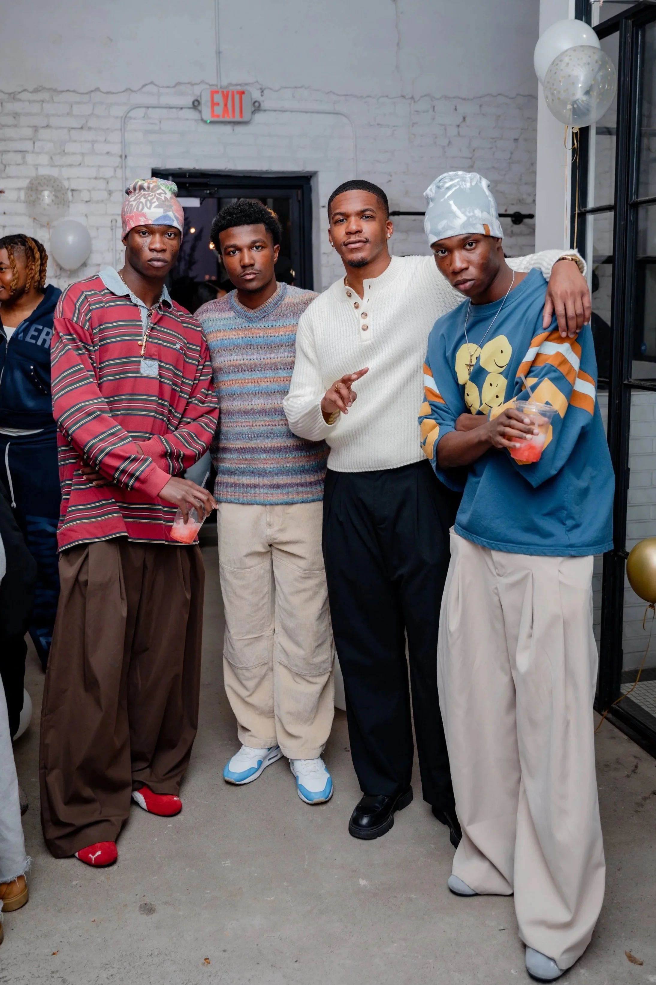 Four young men standing close together at a party, dressed in retro and casual clothing, with balloons and an exit sign in the background.