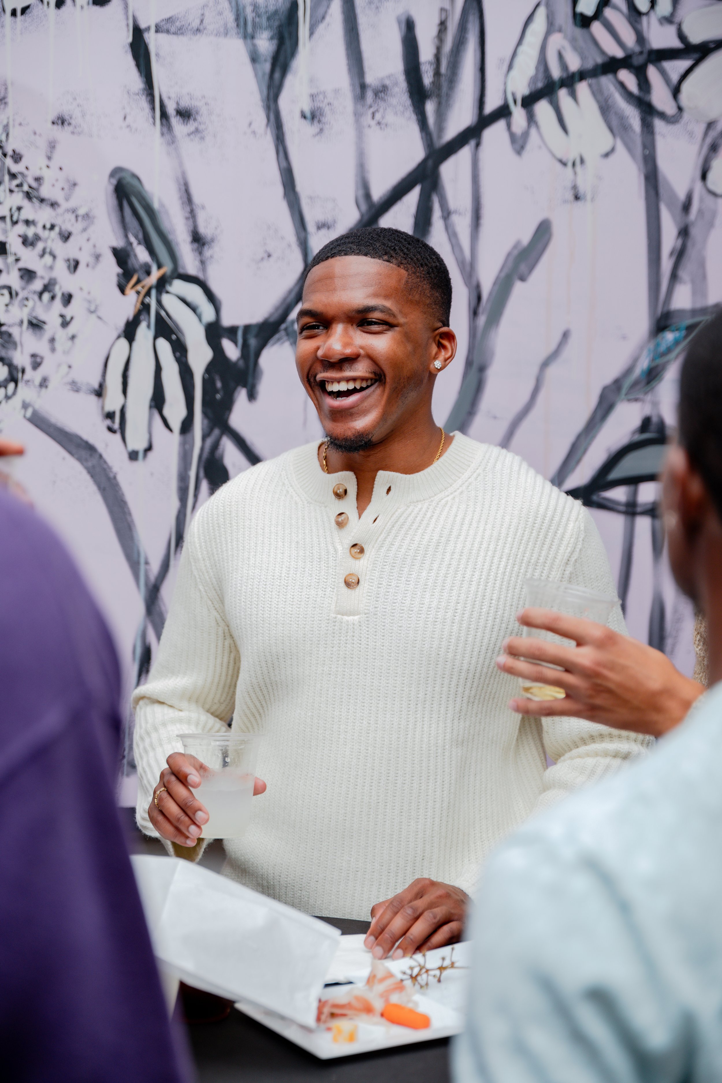 A man with short hair, earrings, and a gold chain, smiling and holding a drink, stands among a group of people at a social gathering. The background has a black and white abstract mural.