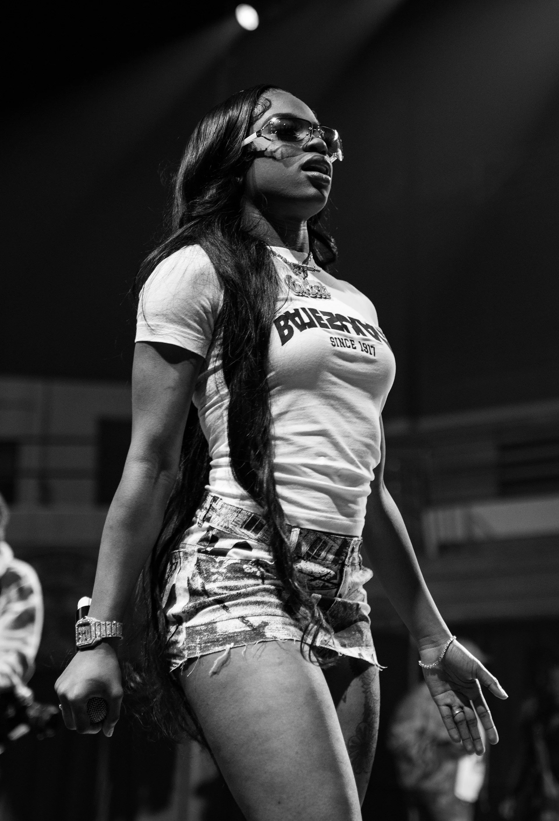 A woman wearing sunglasses, a t-shirt with 'BUZZETTYS SINCE 1917' written on it, and ripped denim shorts, holding a microphone in her right hand, on stage in a black-and-white photo.