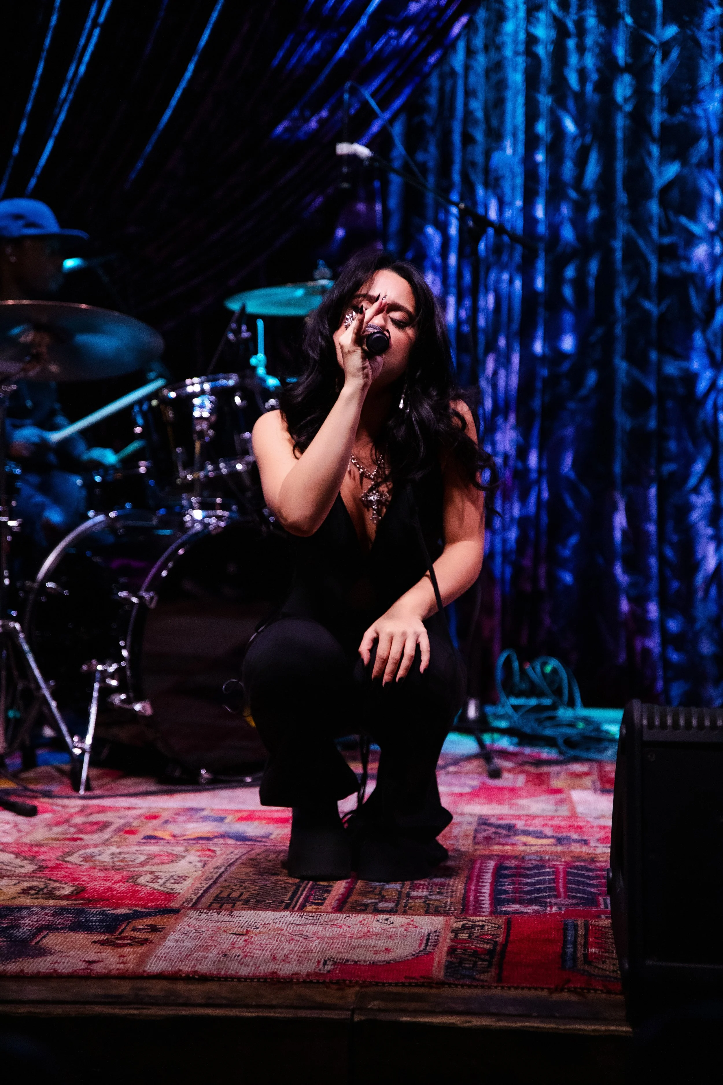 A female singer performing on stage, kneeling with a microphone in her hand, wearing black clothes with dark, wavy hair, with a drummer in the background and colorful stage lighting.