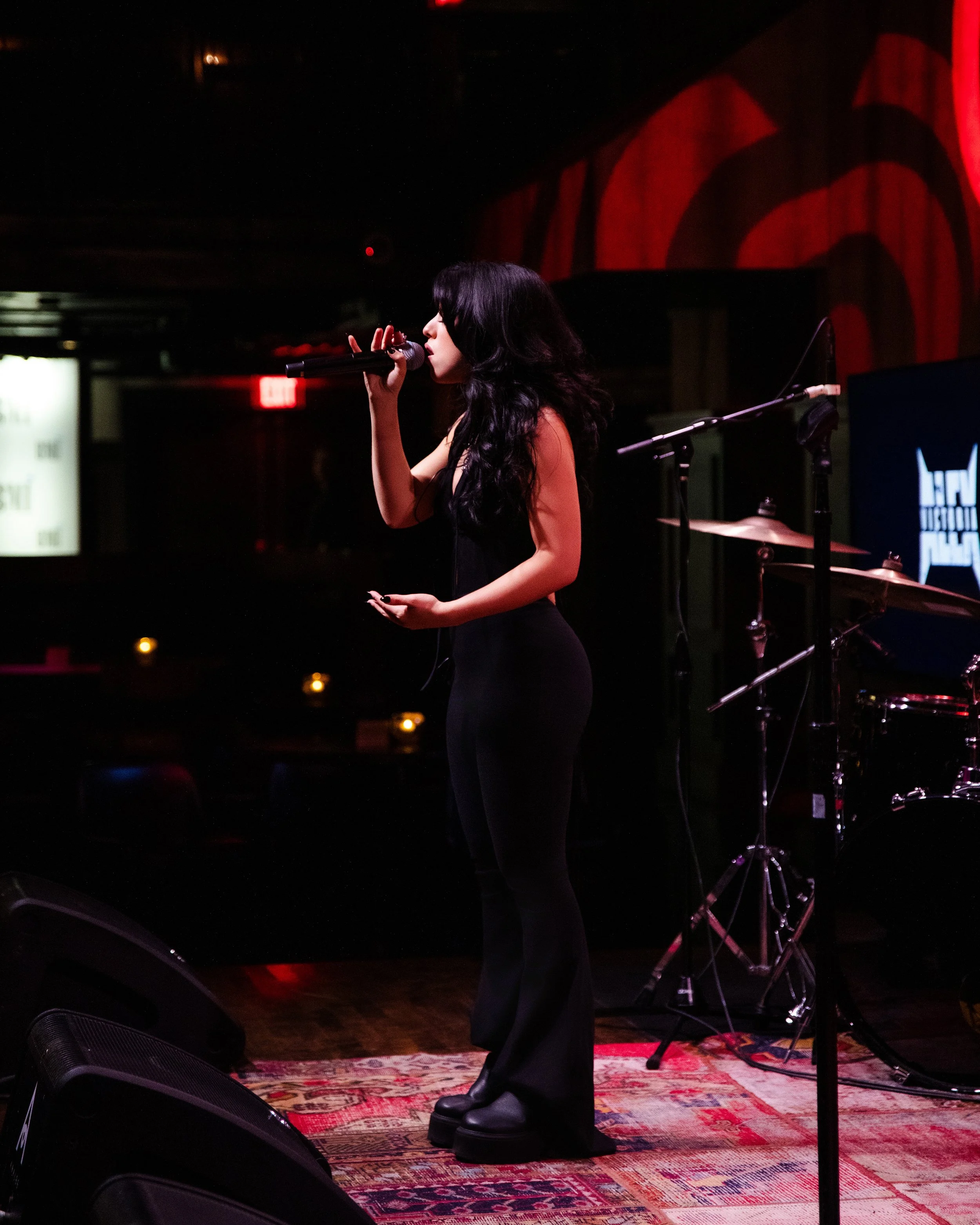 A woman with long black hair singing into a microphone on a dimly lit stage with a drum set behind her.