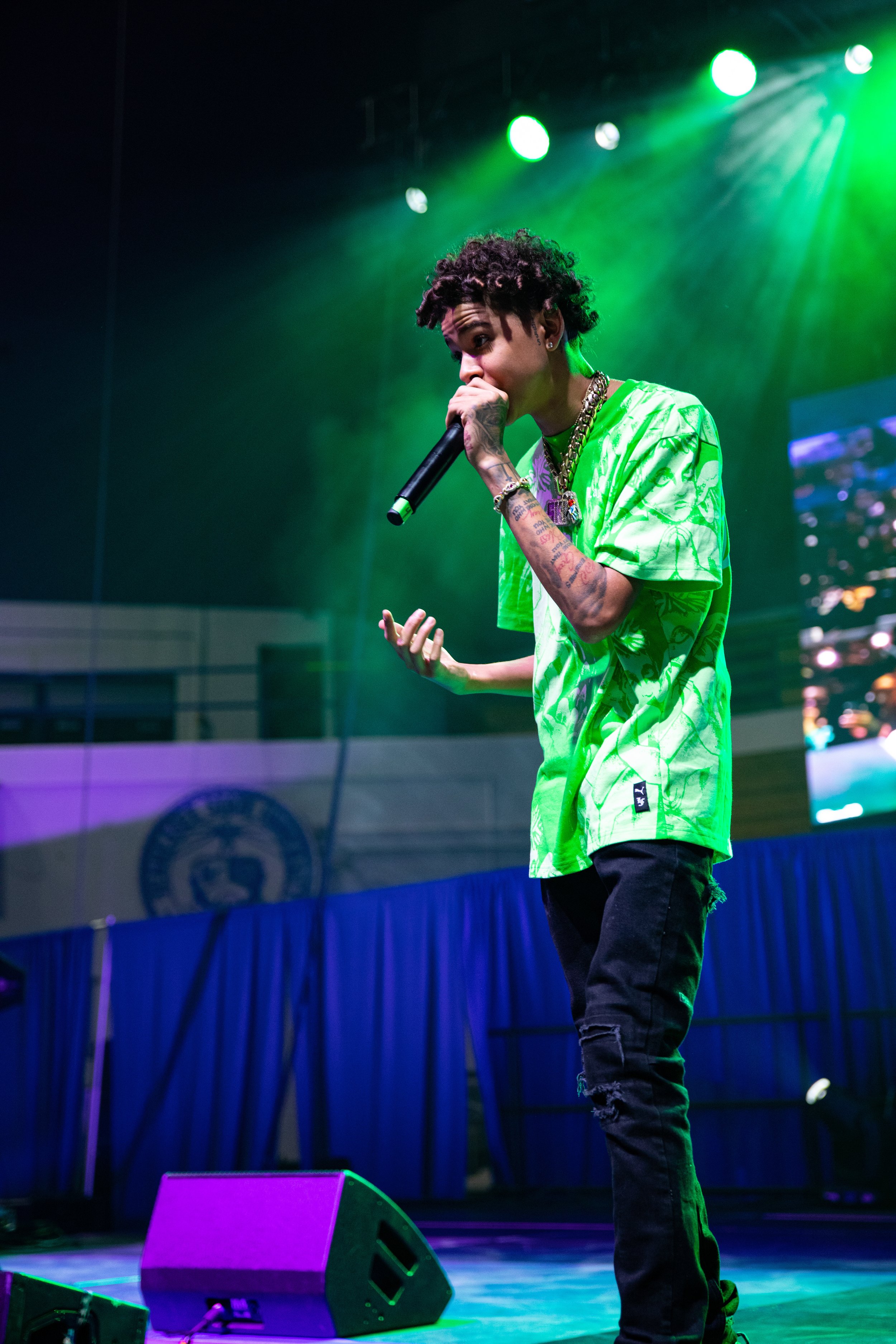 A male performer on stage holding a microphone, wearing a bright green shirt, black ripped jeans, and jewelry, with a green spotlight shining overhead.