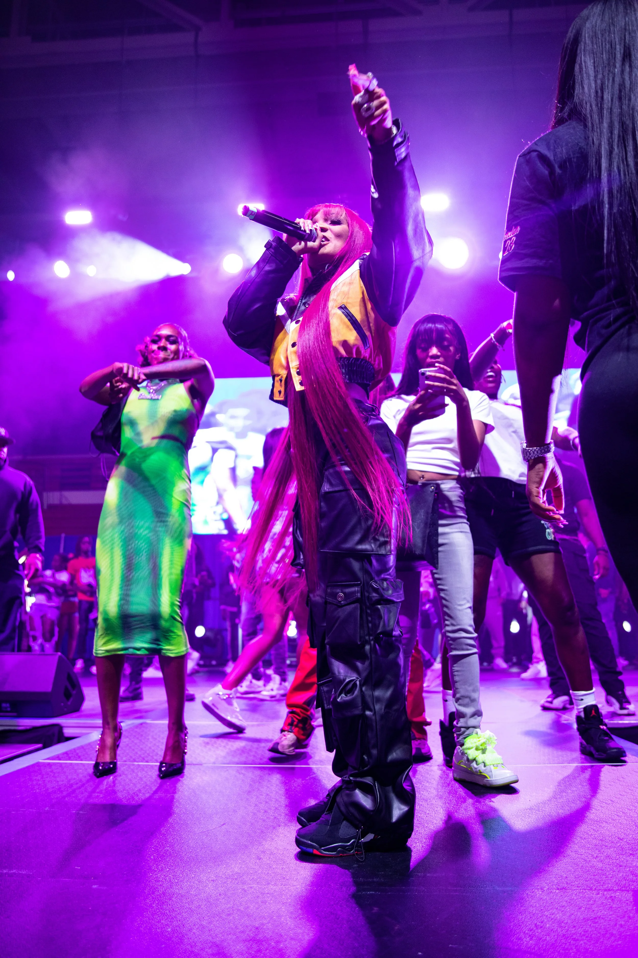 Female performer with long pink hair singing on stage holding a microphone with purple and blue stage lighting, surrounded by people.