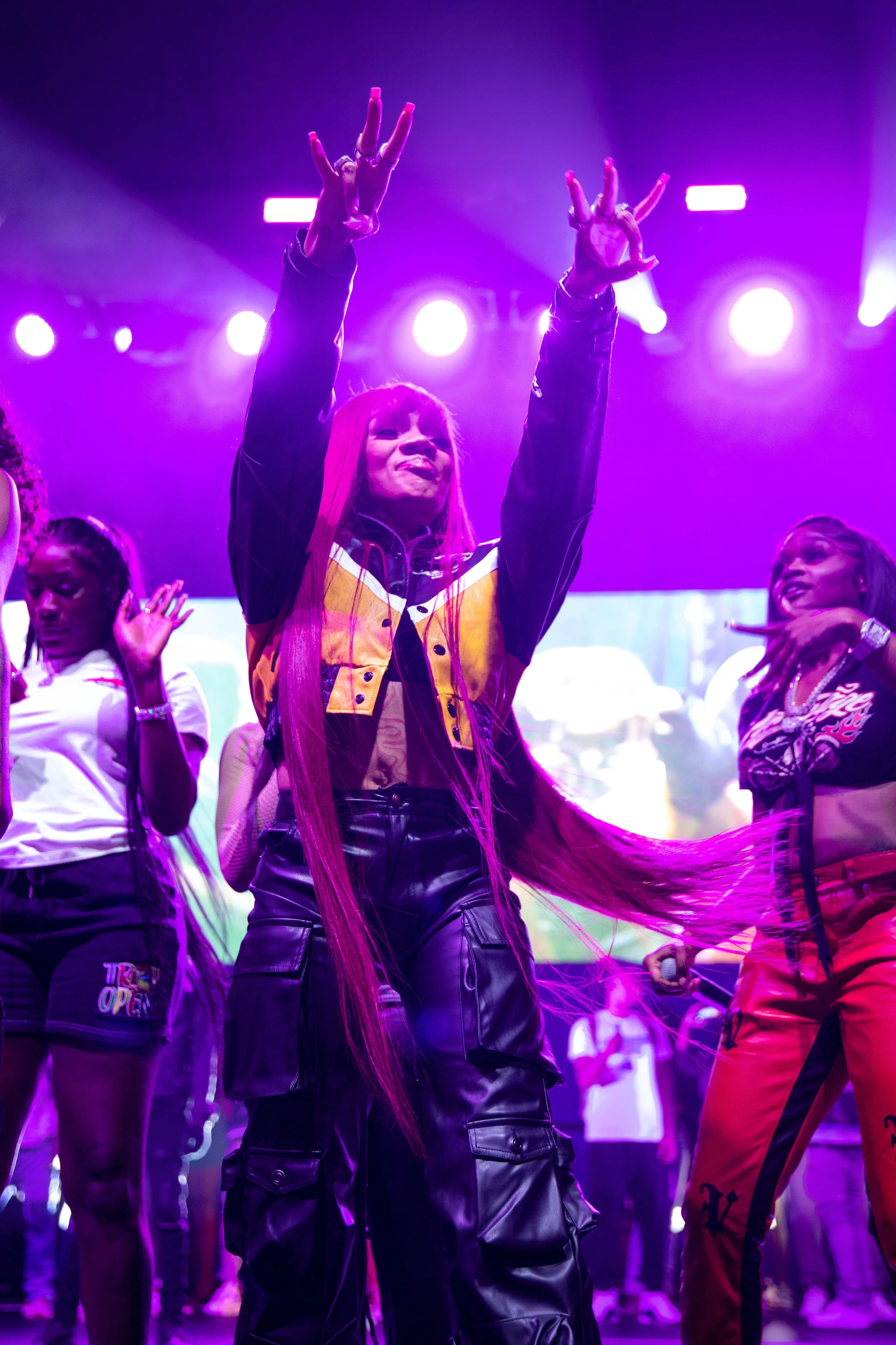 A woman with long pink hair on stage during a performance, surrounded by dancers in colorful outfits, under purple stage lighting.