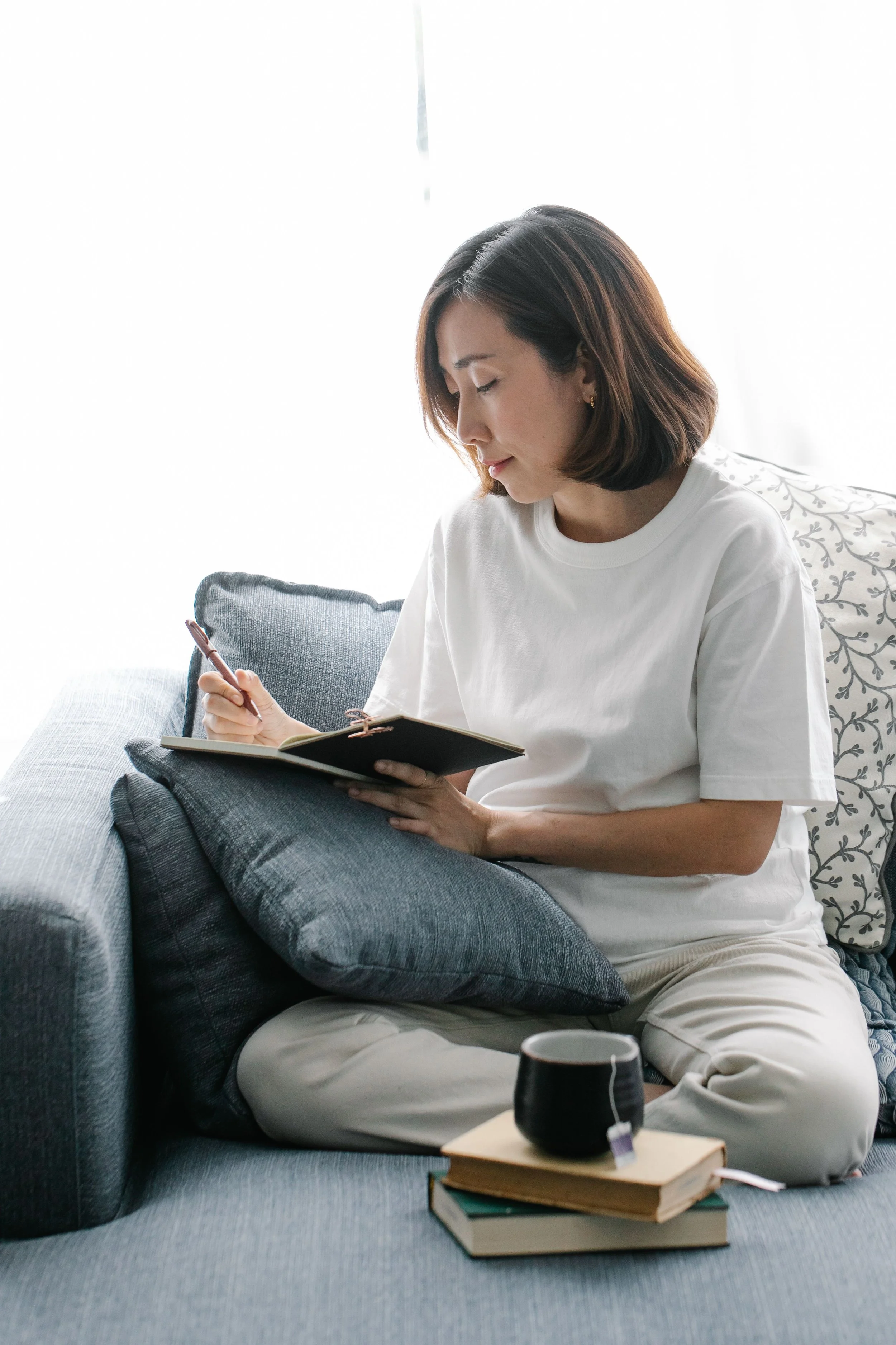 A woman with shoulder-length brown hair wearing a white T-shirt, sitting on a grey couch with dark grey pillows, and reading or writing in a notebook, with a stack of books and a black cup on the table in front of her, in a room with a bright window behind her.