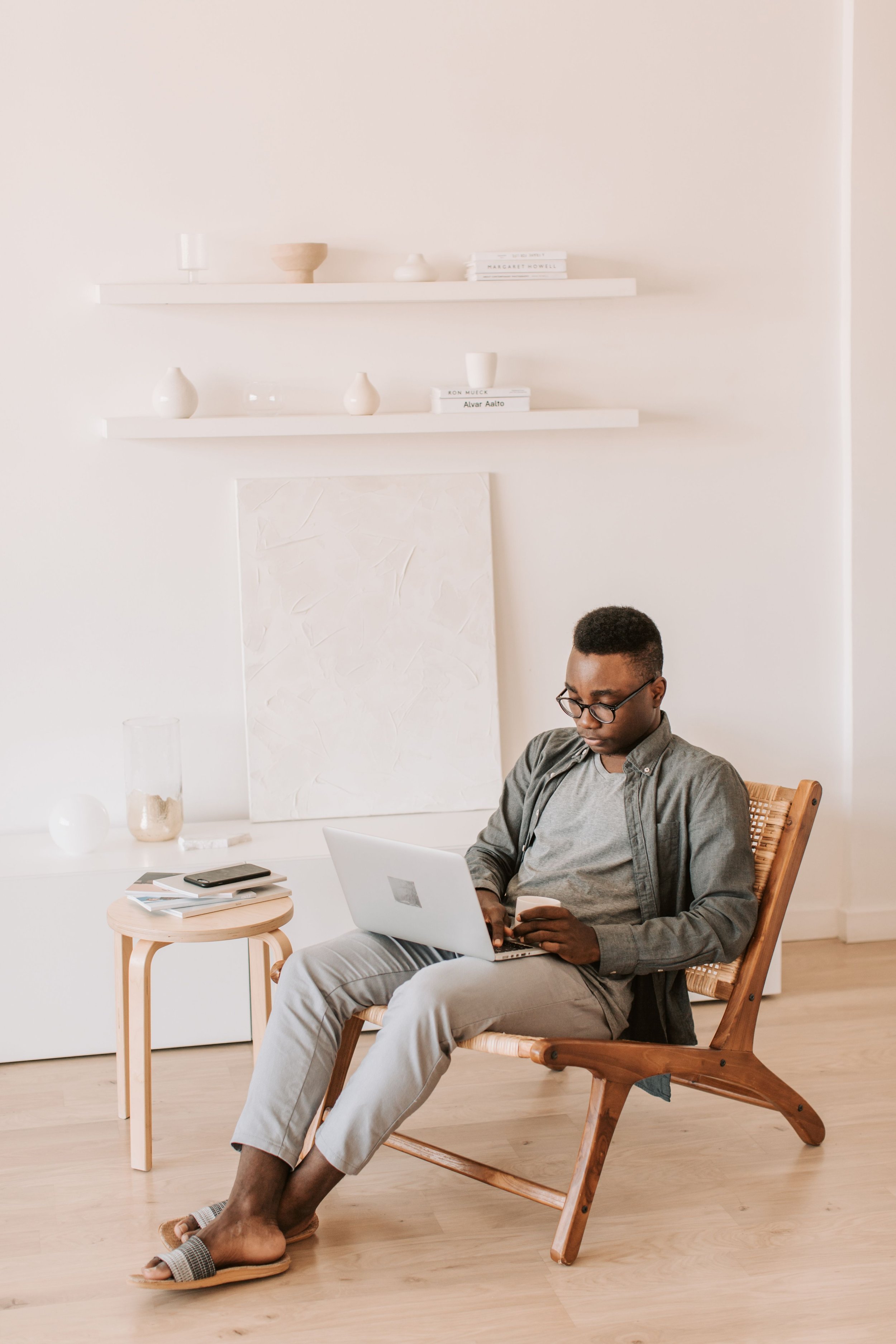 A man seated in a wooden chair, using a laptop on his lap, in a minimalist, bright room with white walls and floating shelves holding decorative items and books.