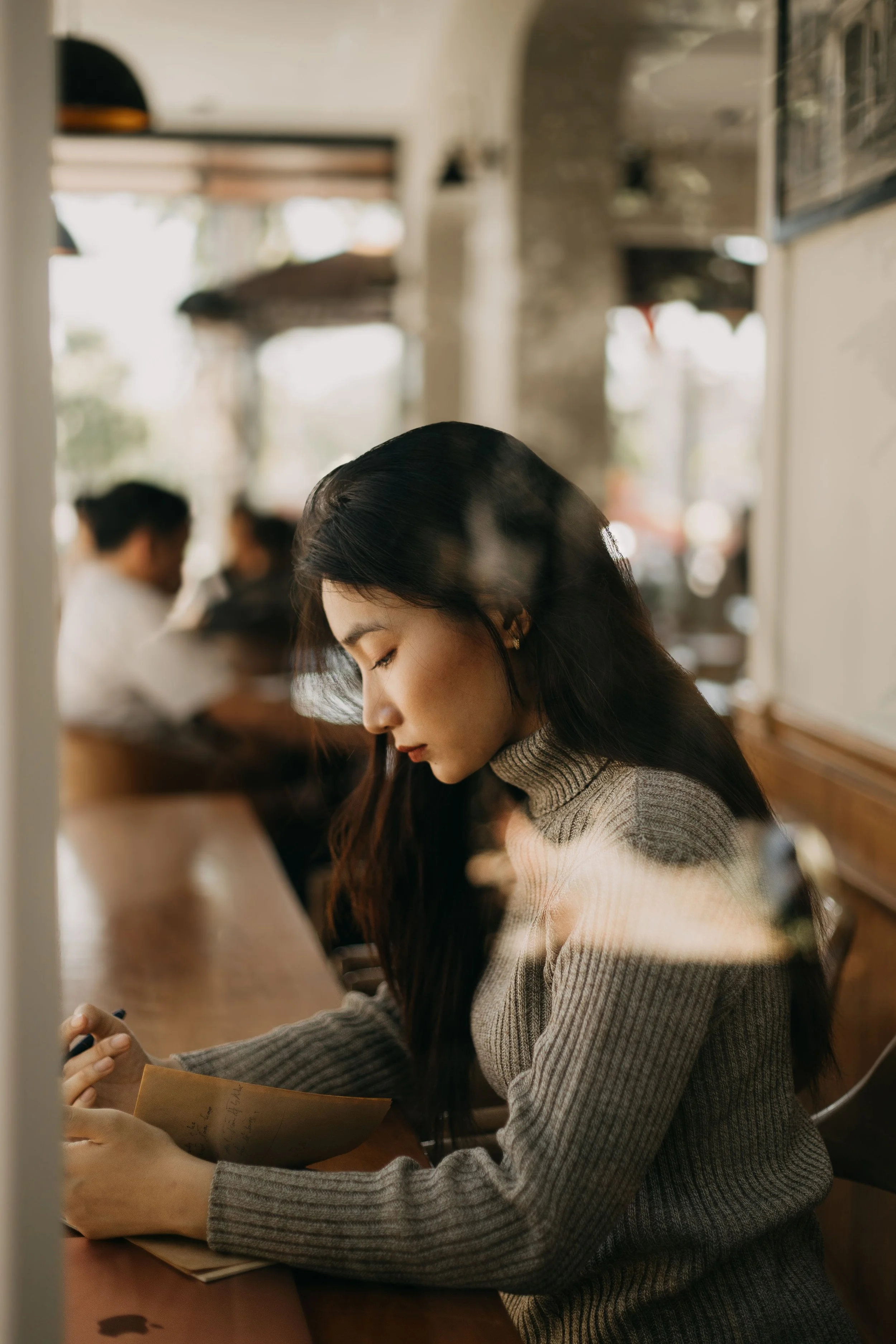 A young woman with long dark hair and a gray turtleneck sweater sitting at a table in a cafe or restaurant, writing or reading a paper, with a blurred background of other patrons and large windows with natural light.