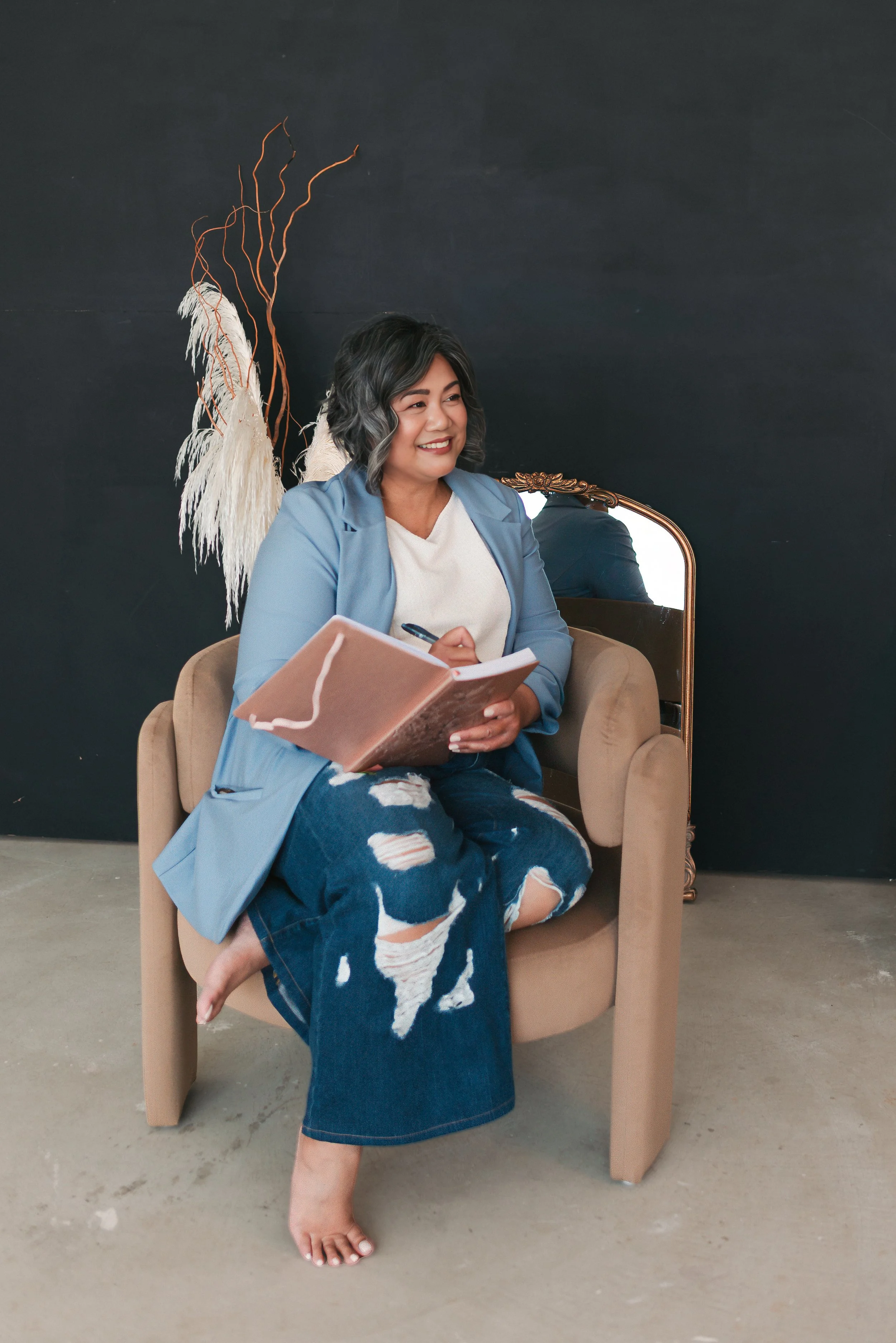 Edmonton therapist sitting barefoot in a beige armchair, smiling and holding an open notebook with a pen, wearing a white t-shirt, blue blazer, and ripped jeans, with a decorative feathered and plant arrangement behind her against a dark wall.