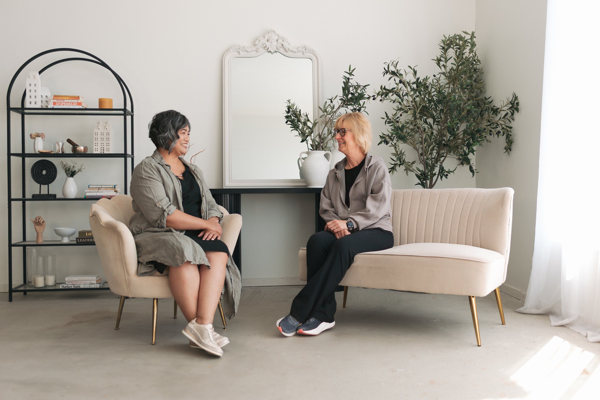 Two women are sitting and talking in a bright living room, smiling at each other. One woman has short curly gray hair and is wearing glasses, a gray hoodie, and black pants. The other woman has short dark hair with gray streaks, wearing a tan coat over a black top, and light-colored sneakers. Behind them is a large decorative mirror, a white couch, a black shelving unit with books and decorative items, and a large plant.
