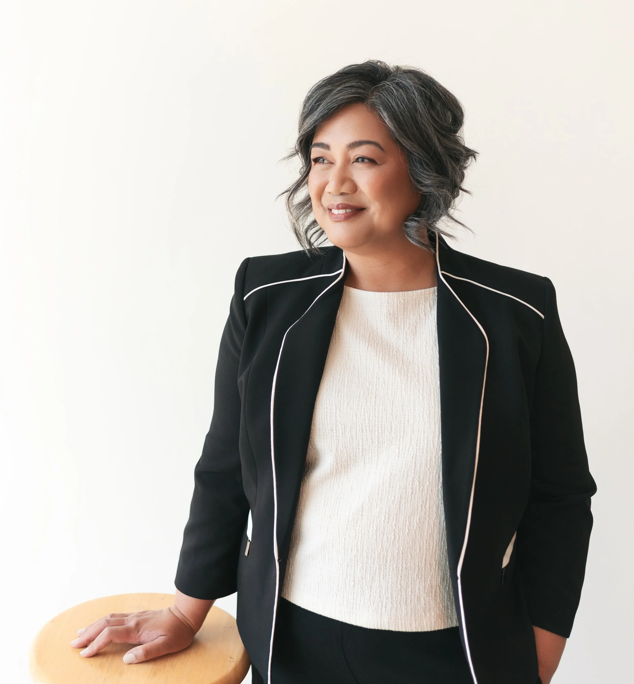 Rhoda Lalog, counselling therapist in Edmonton. A woman with short, gray hair, wearing a black blazer with white piping and a cream-colored top, resting her hand on a wooden table.