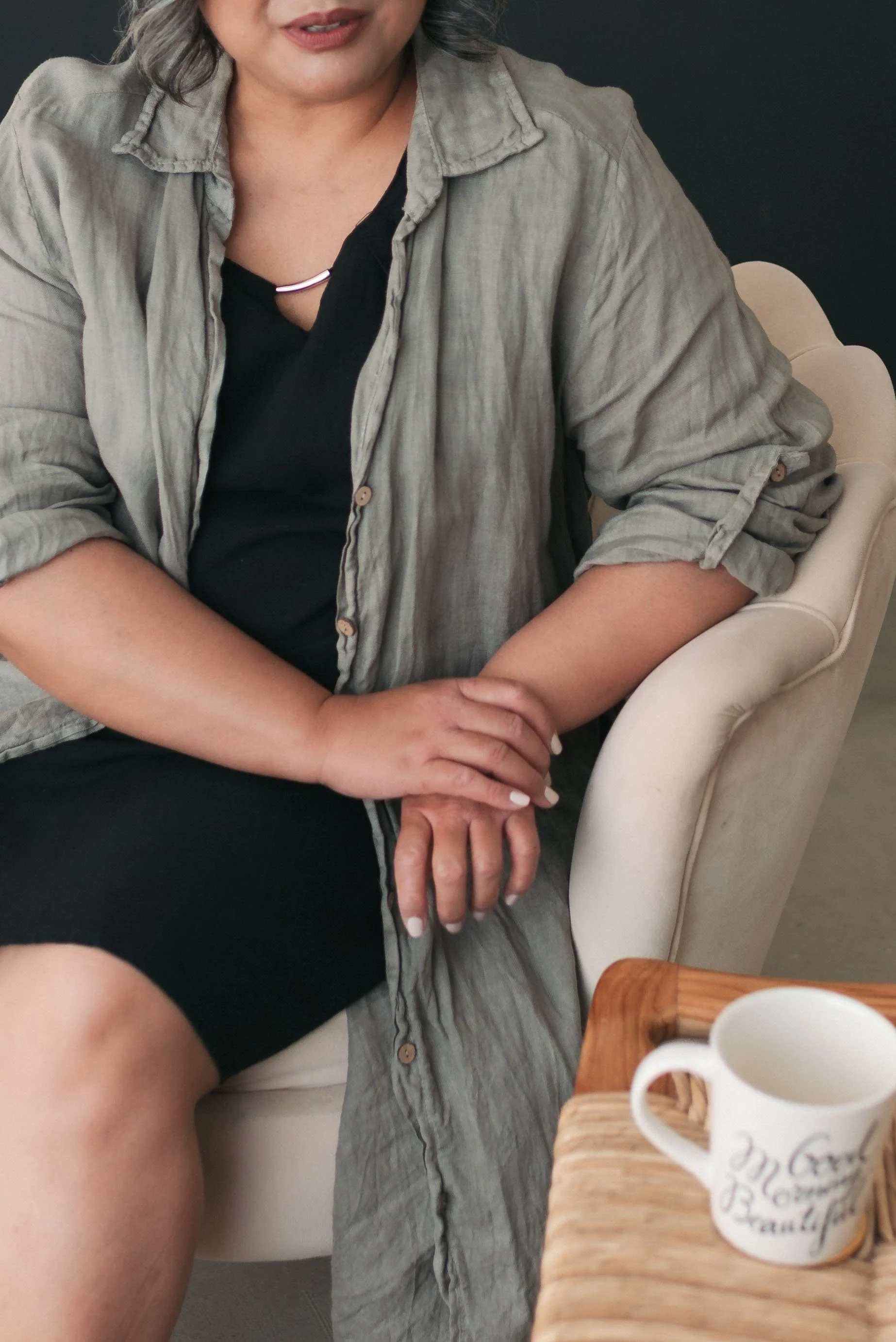 A woman sitting on a beige chair wearing a gray button-up shirt over a black top, with a silver necklace, and her hands clasped in her lap. There is a wooden table with a white mug in the foreground.