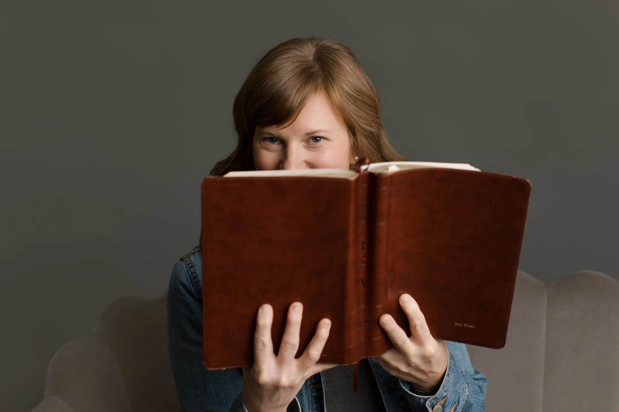 A woman with brown hair holding a large brown hardcover book, partially hiding her face, looking at the camera, against a plain gray background.