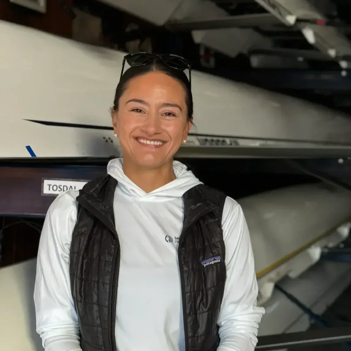 Smiling woman with sunglasses on her head, wearing a white Patagonia jacket and black vest, standing in front of rowing shells stored in a boathouse.