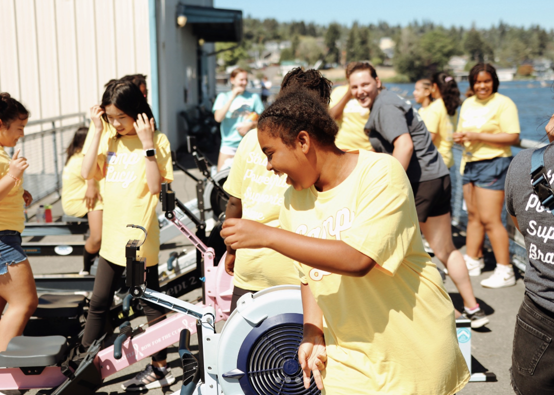 Group of smiling women and girls in yellow t-shirts gather outdoors near water, with some using rowing machines on a dock on a sunny day.