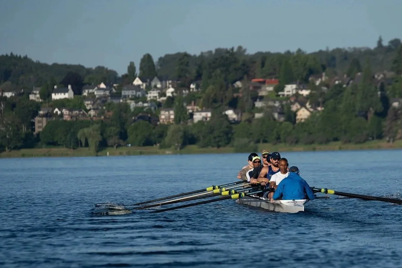 A group of people rowing a boat on a lake with houses and trees in the background.
