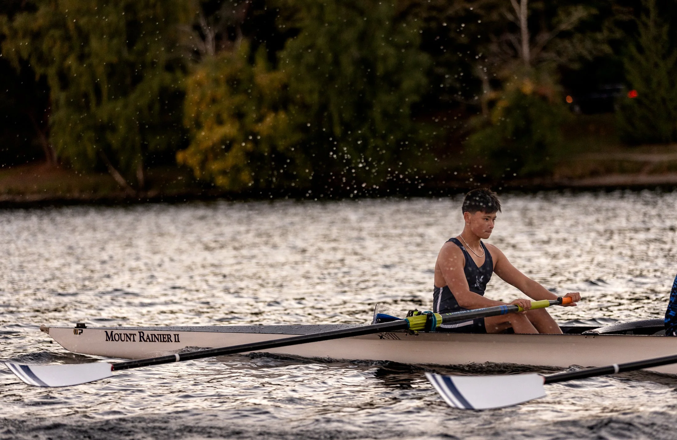 A young rower on a rowing boat with the words 'Mount Rainier II', sitting on a river during dusk or dawn, splashing water as they row, with trees in the background.