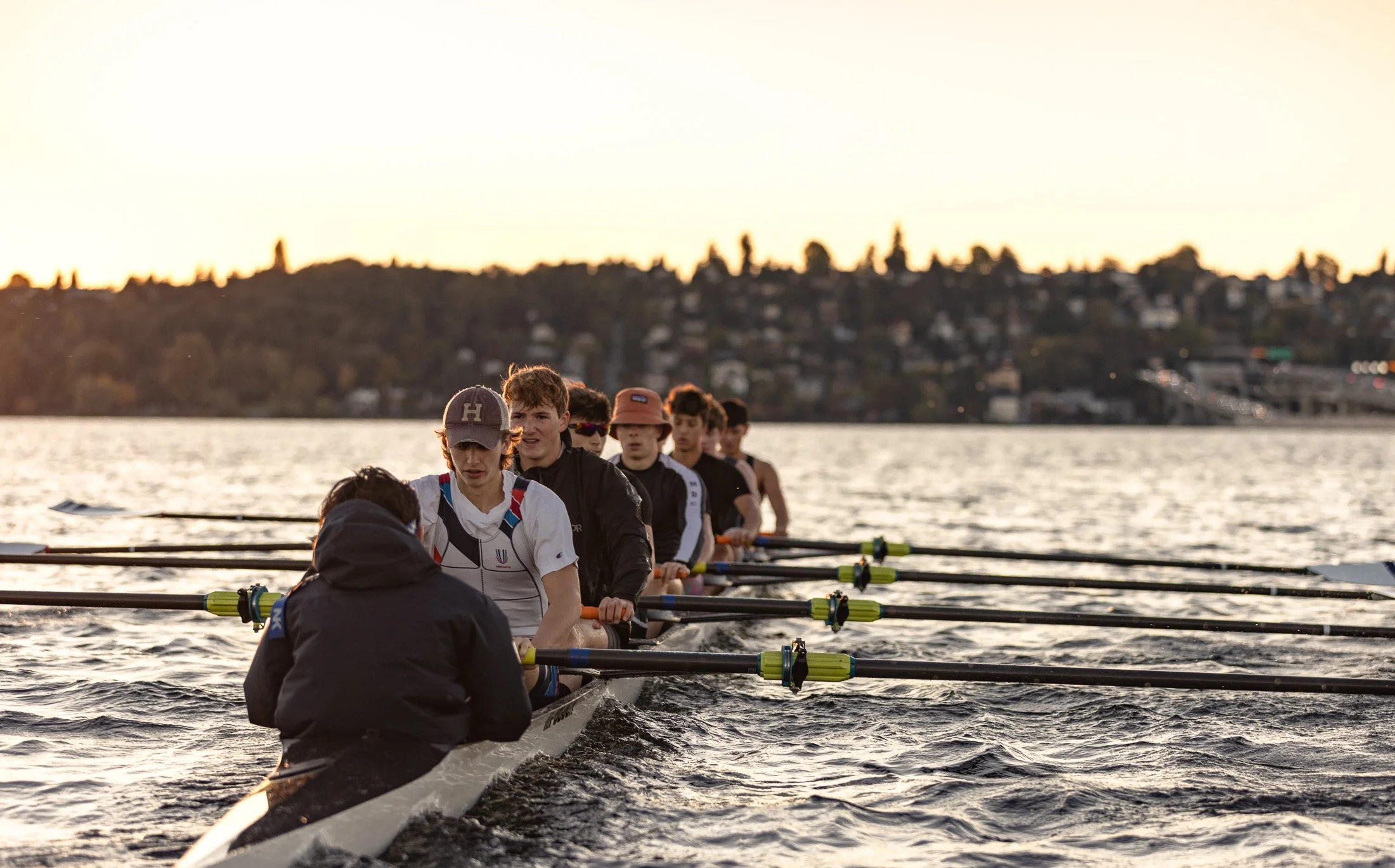 A group of young rowers in a racing shell on the water at sunset, with a cityscape in the background.