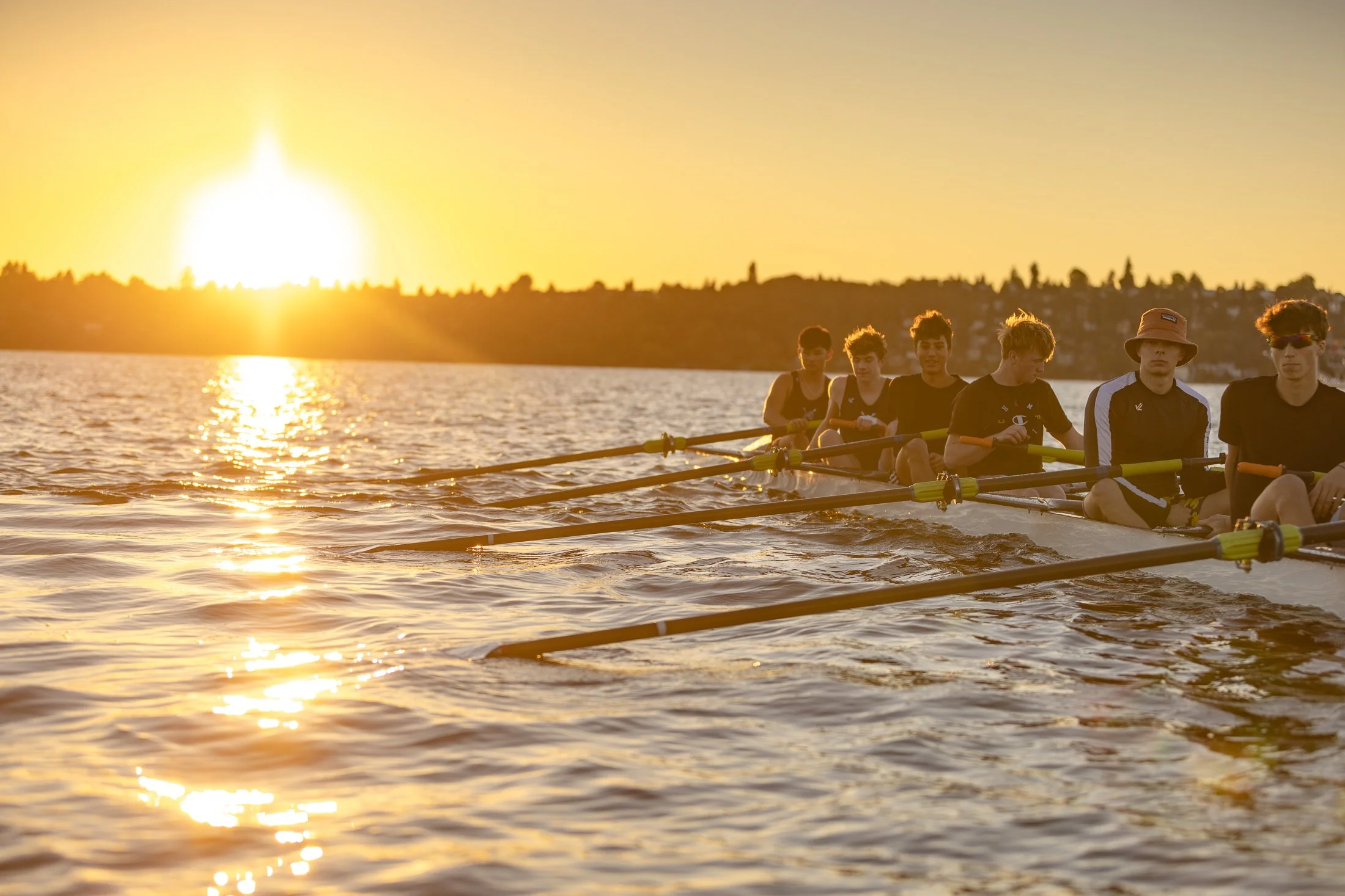 A group of young men rowing a racing boat on a lake during sunset.