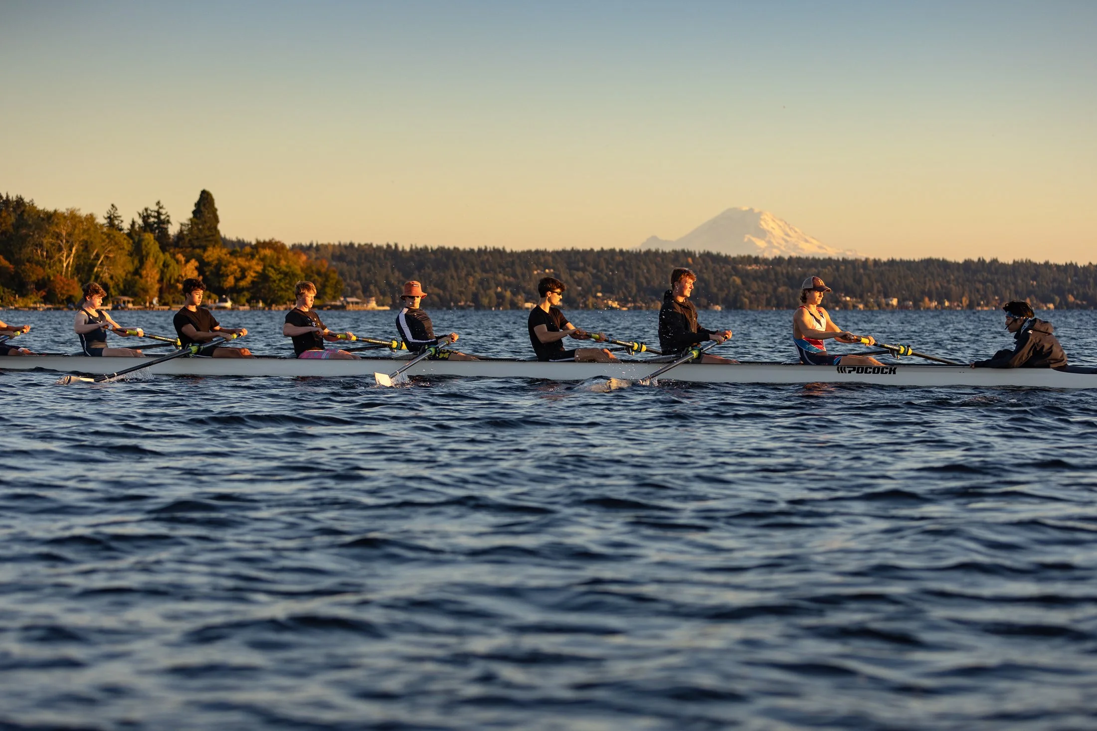 A group of rowers on a boat racing on a lake during sunset, with trees and a snow-capped mountain in the background.