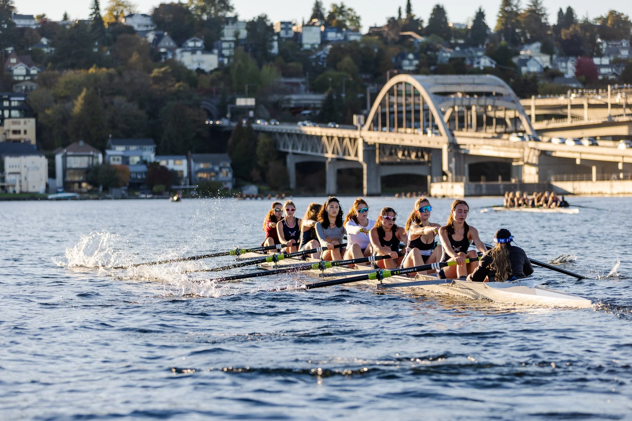 A team of women in a rowing boat competing on a river during sunset, with a city bridge and hillside residential area in the background.