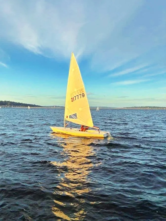 A person sailing a small yellow sailboat on a calm body of water during late afternoon or early evening, with a partly cloudy sky and distant land visible on the horizon.