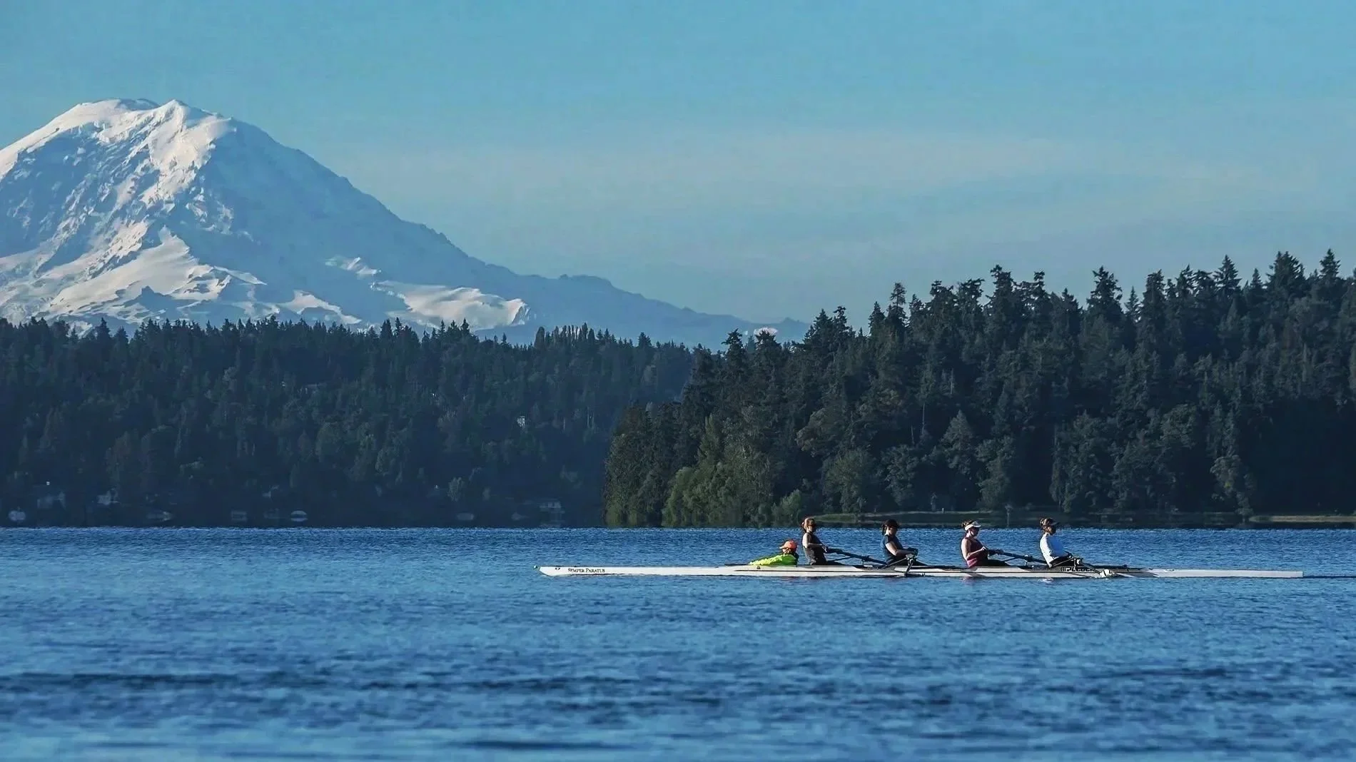 A group of four people rowing a boat on a calm lake with a forest and a snow-capped mountain in the background.