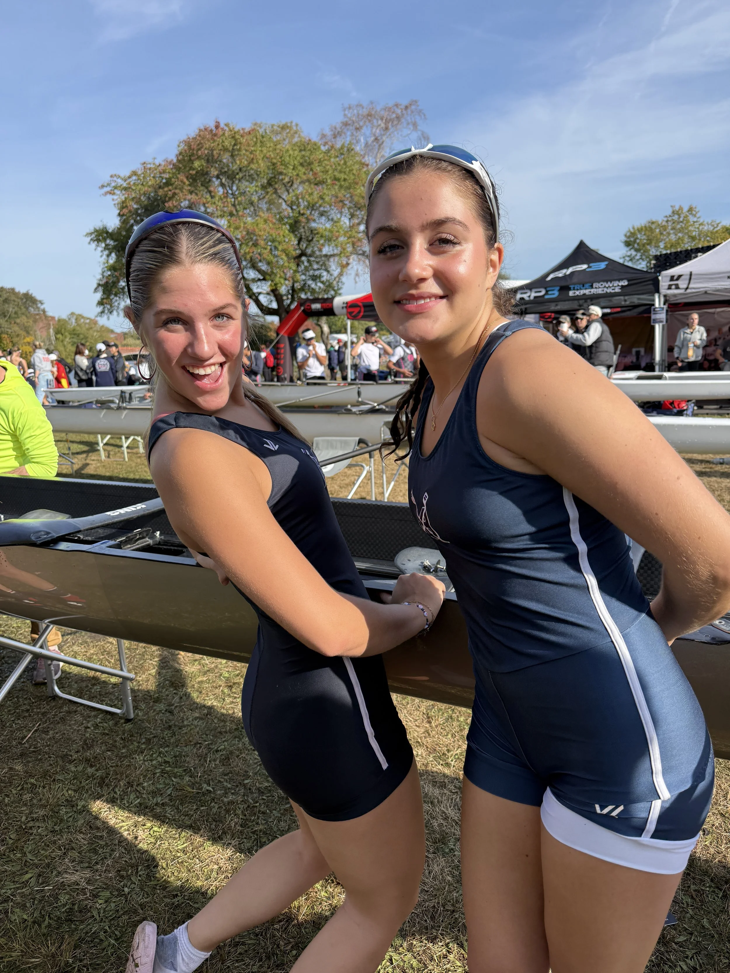 Two young women in athletic uniforms smile and pose together at an outdoor rowing event, with rowing shells and event tents in the background.