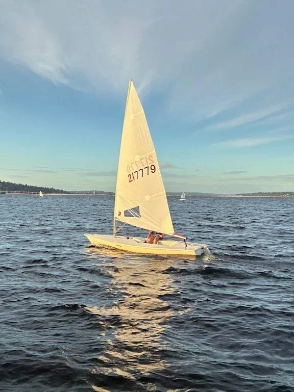 A small sailboat with white sails on a calm body of water under a blue sky, with a distant shoreline and a few other boats.