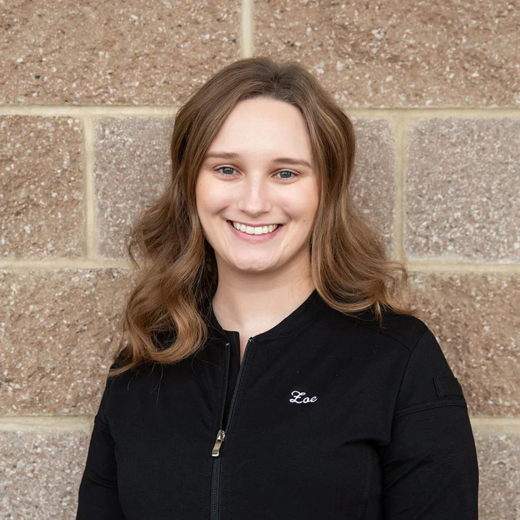 A massage therapist standing in front of a brick wall. She is wearing a black jacket with the name 'Zoe' embroidered on it.