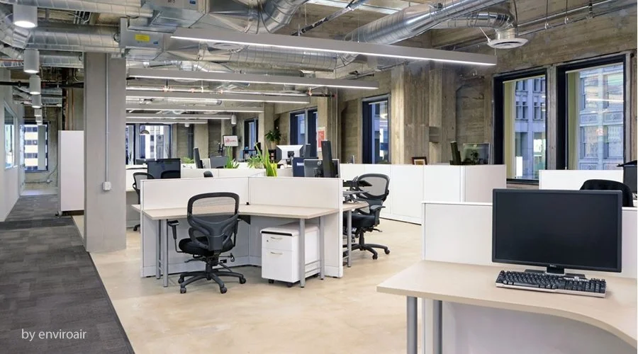 Modern open-plan office with workstations, chairs, and computer monitors, large windows, exposed ceiling ducts, and a combination of carpeted and concrete flooring.