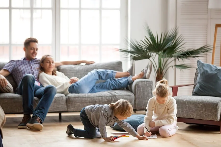 A family relaxing in a bright living room with large windows, a man and a woman on a gray sofa, and two children playing on the floor.