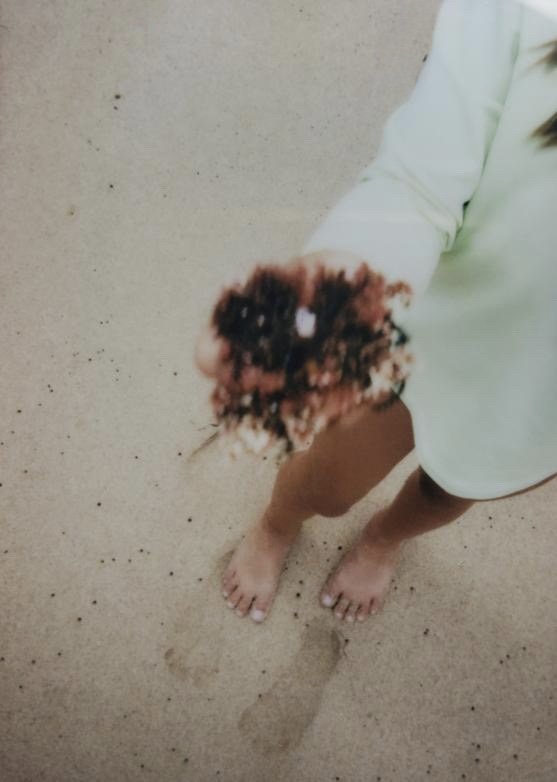 A photograph of a person in a teal shirt on a sandy beach, holding a coral’s, with footprints on the sand visible below.