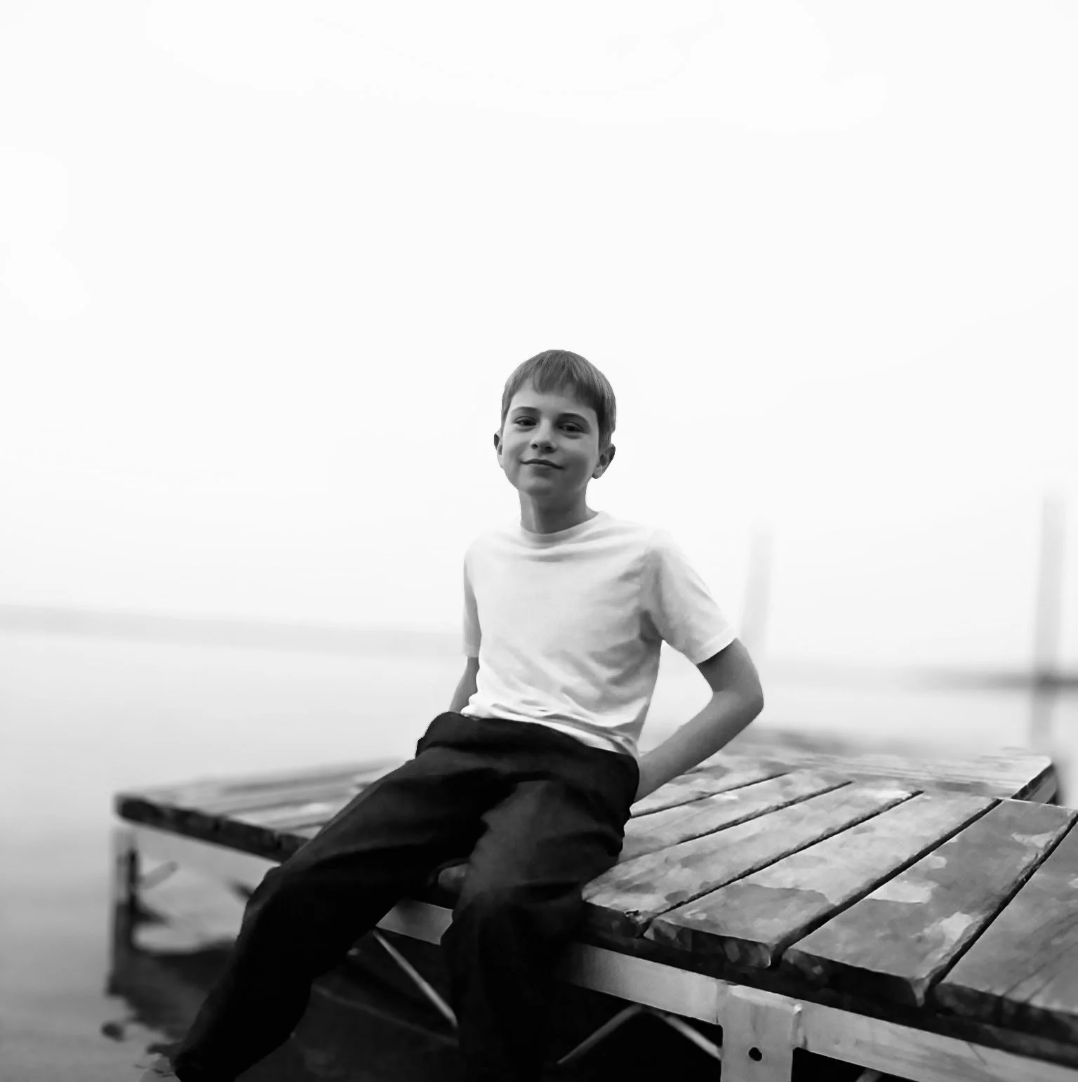 A young boy in a white T-shirt and dark pants sitting on a wooden bench outdoors, smiling at the camera in black and white.