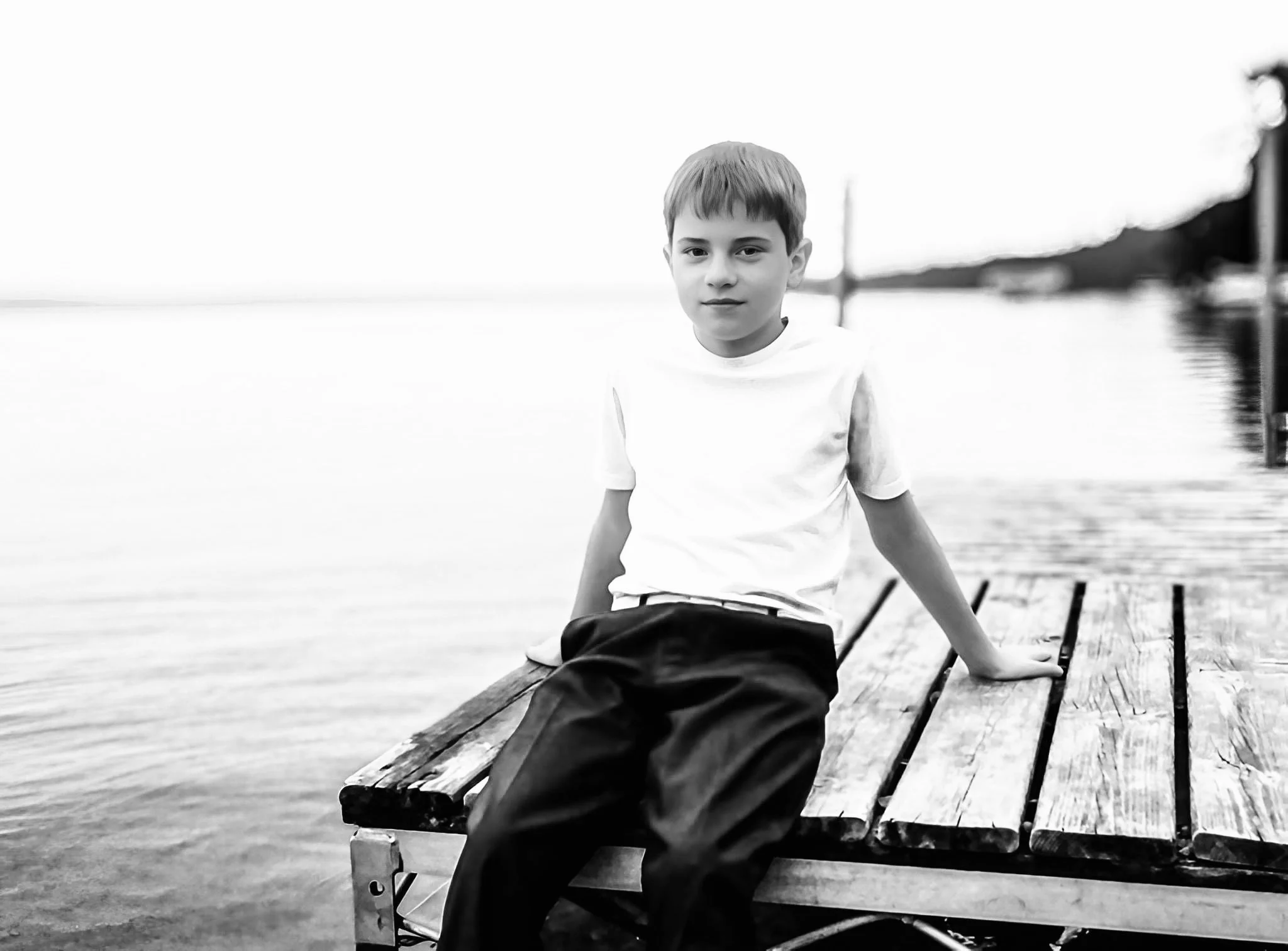 A young boy with short hair sitting on a wooden dock by the water, looking at the camera, in black and white.