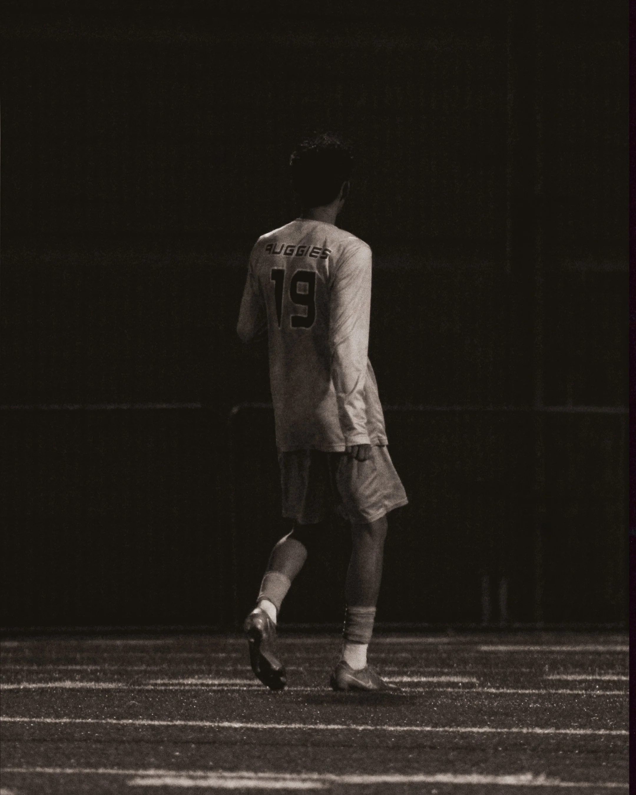A football player in uniform walking on a field at night, facing away, with the name 'Auggies' and number 19 on their jersey.