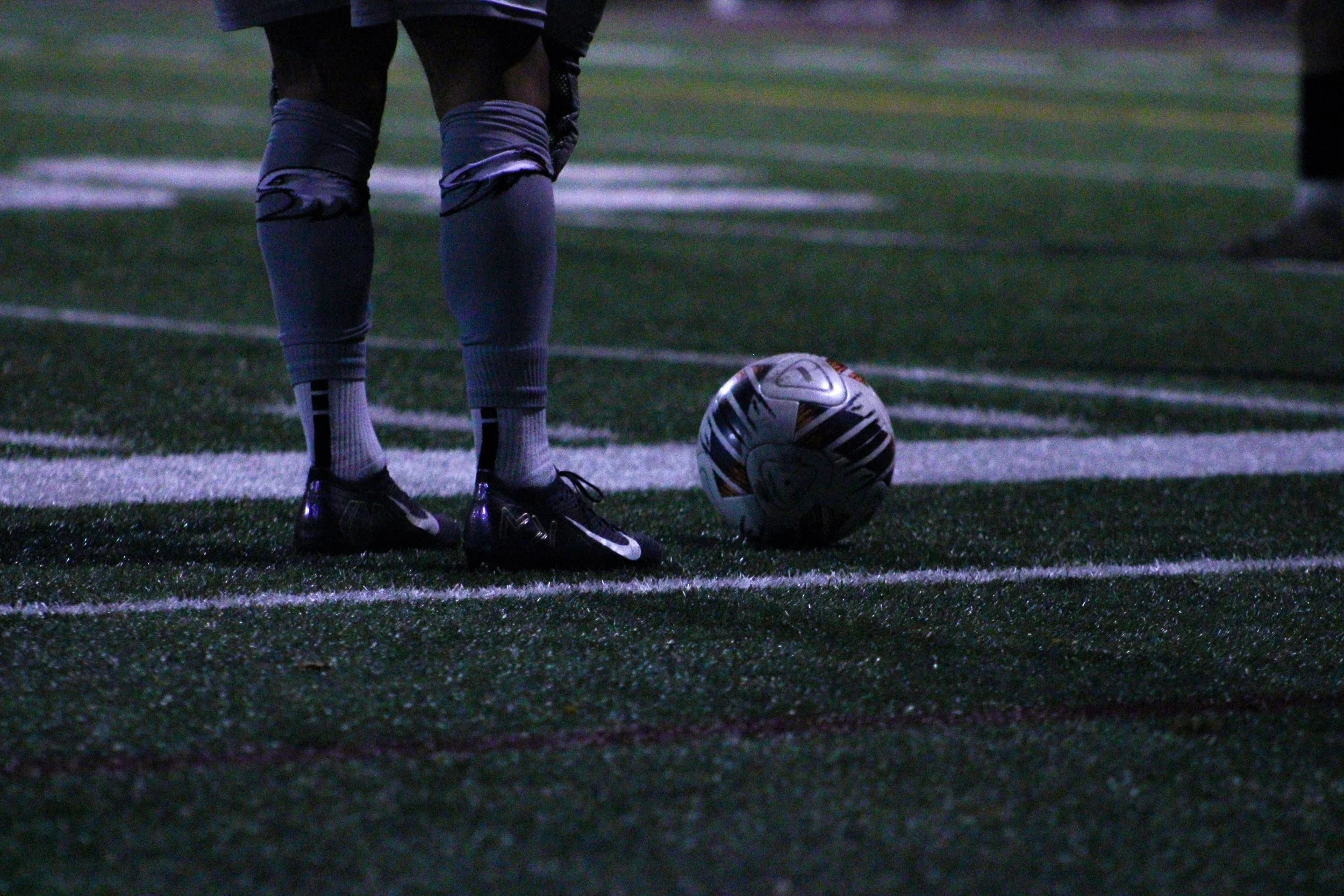 A soccer player standing on a field with their feet beside a soccer ball at dusk or nighttime.