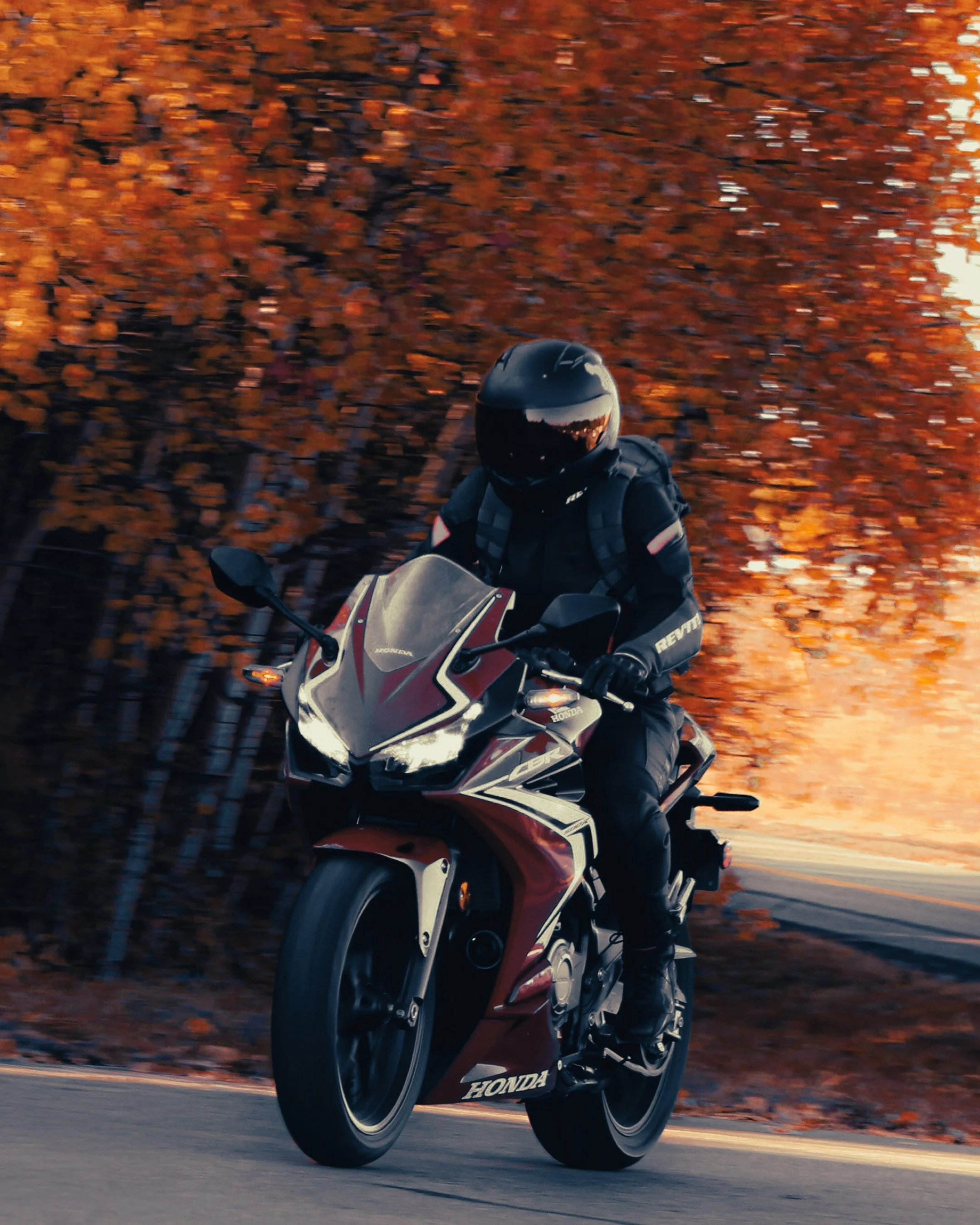 Motorcycle rider wearing a black helmet and gear riding a red and white Honda bike on an outdoor road with autumn foliage in the background.