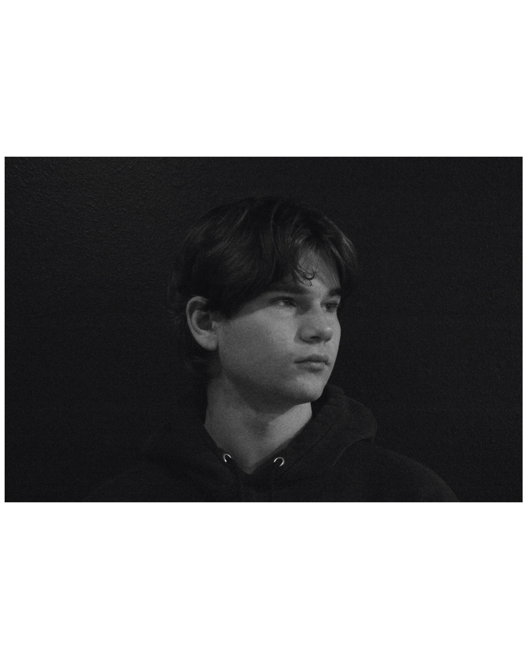 Black and white portrait of a young man with medium-length dark hair, wearing a hoodie, against a dark background.