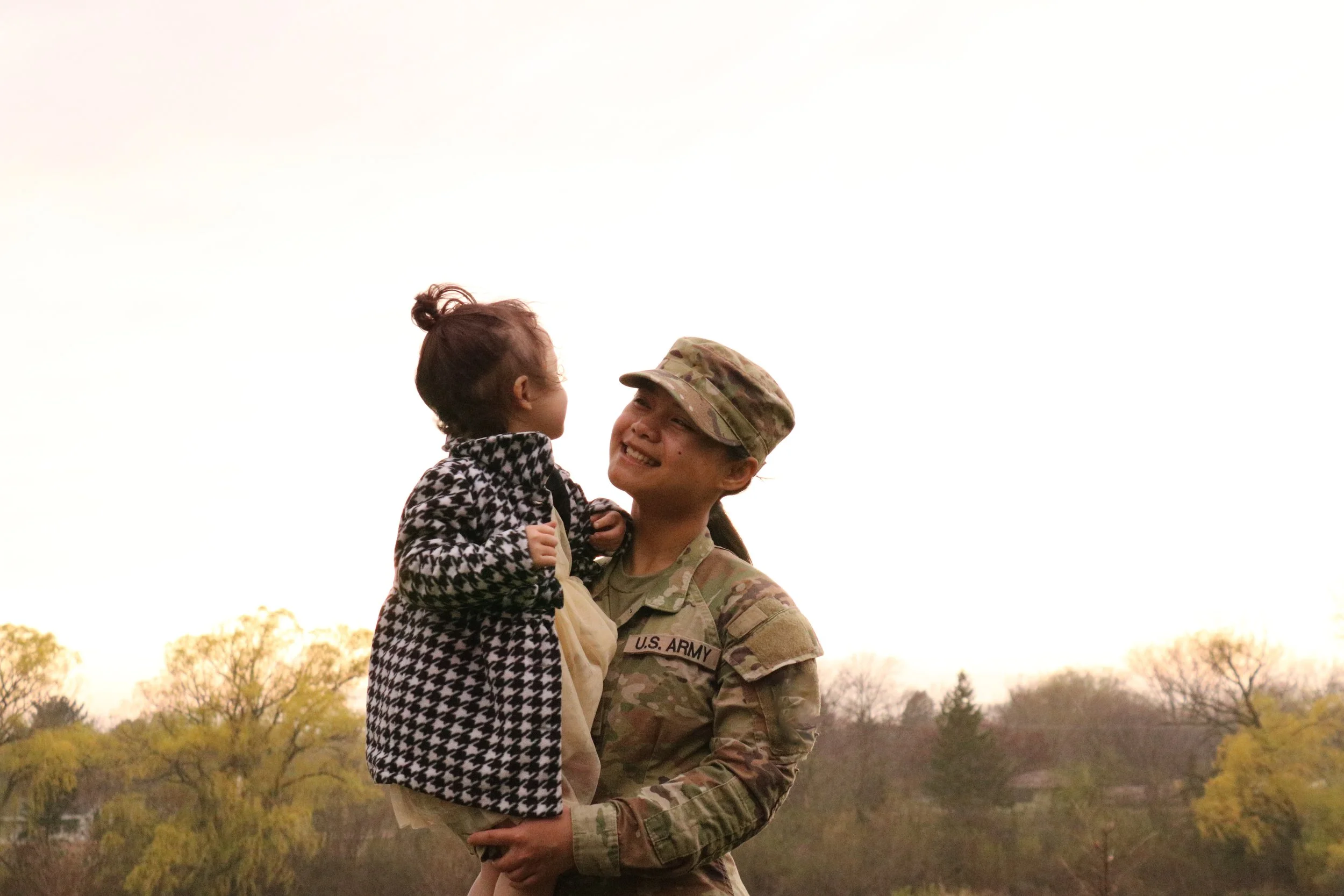 A U.S. Army soldier in uniform holding a young girl in her arms, both smiling and looking at each other outdoors during daytime, with trees and a cloudy sky in the background.
