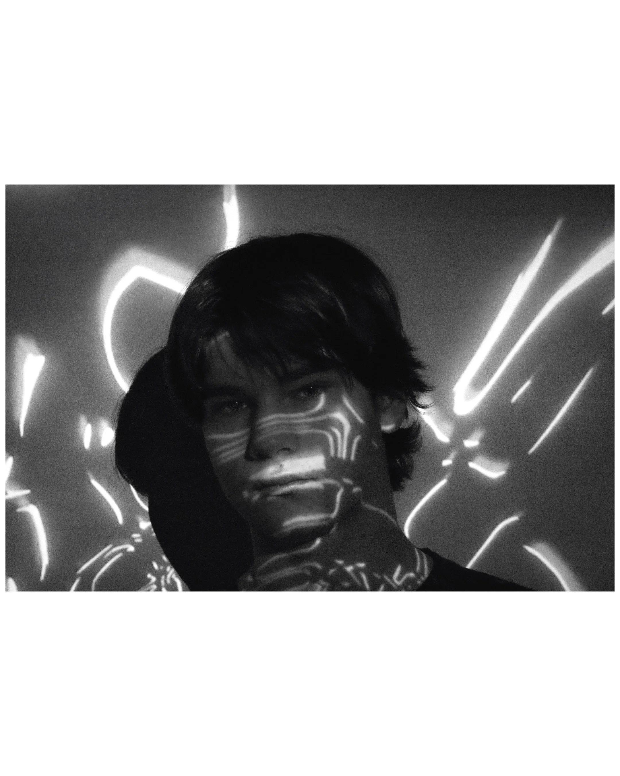Black and white photo of a young man with dark hair, illuminated by abstract light patterns projected onto his face and background.