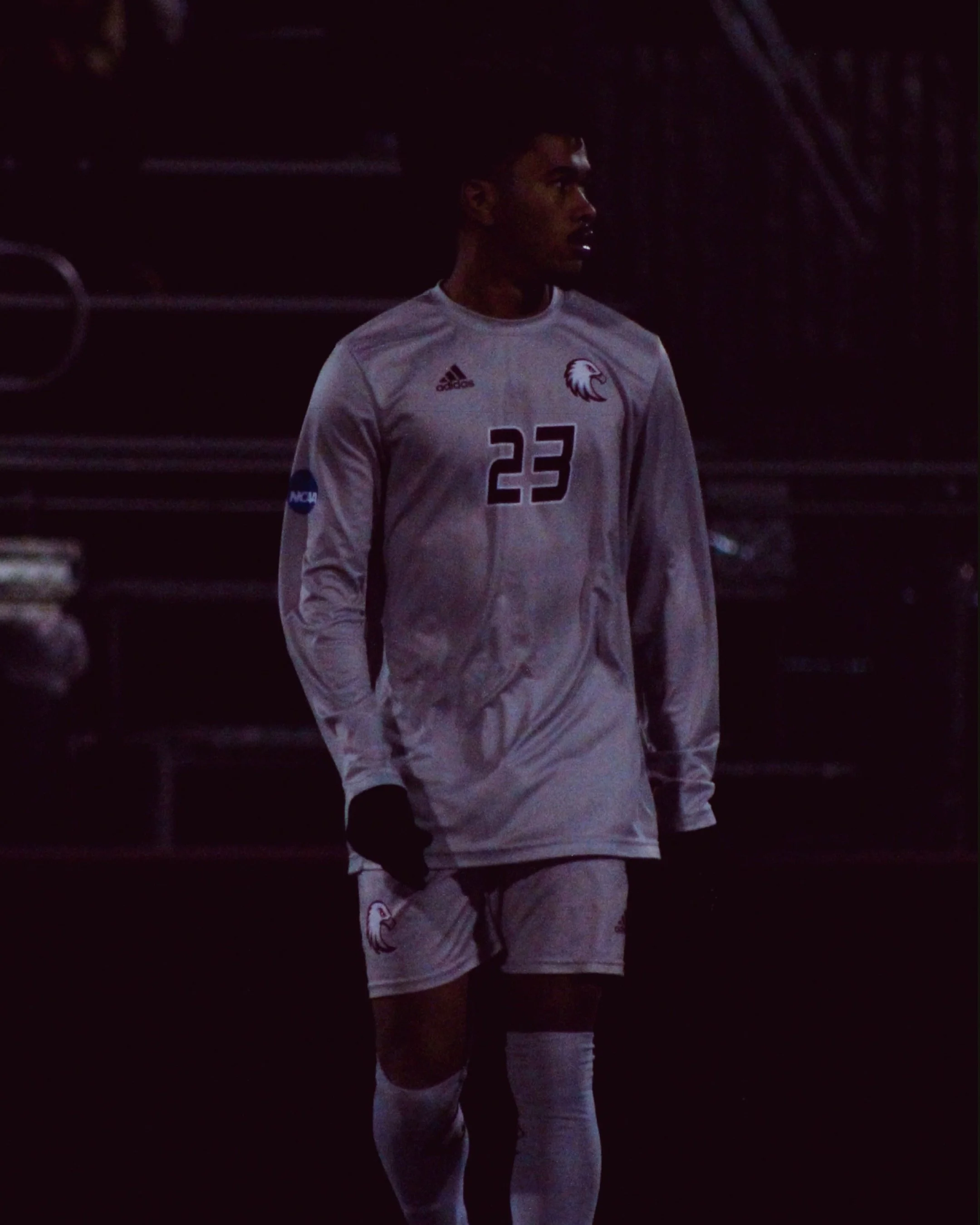 A male soccer player in a beige uniform standing in a dark environment. He is wearing a jersey with the number 23, an Adidas logo, an Eagle logo, and an NCAA patch on the sleeve, along with matching shorts, socks, and black gloves.