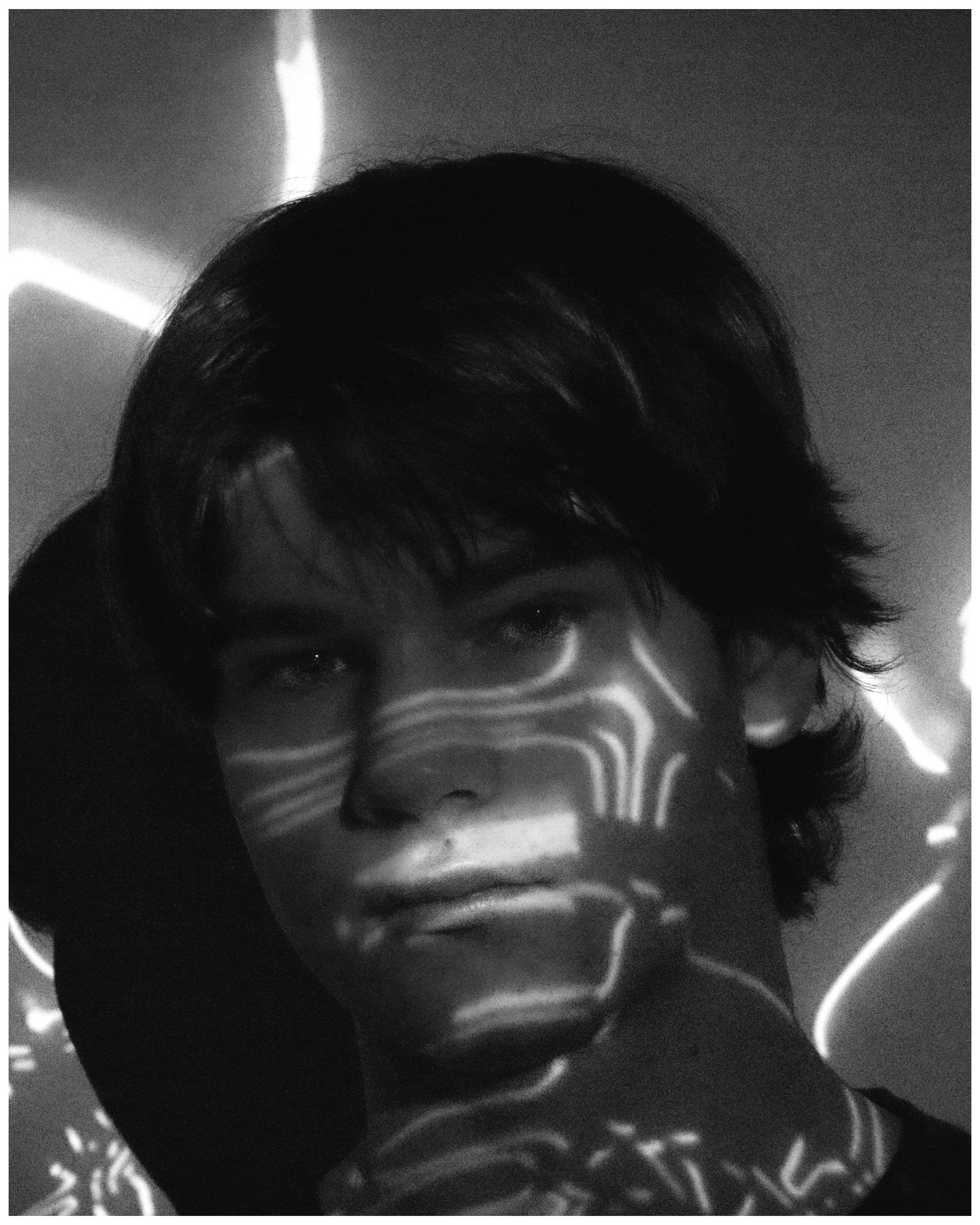 A young person with dark, wavy hair looking at the camera in a dark room with light patterns projected onto their face.