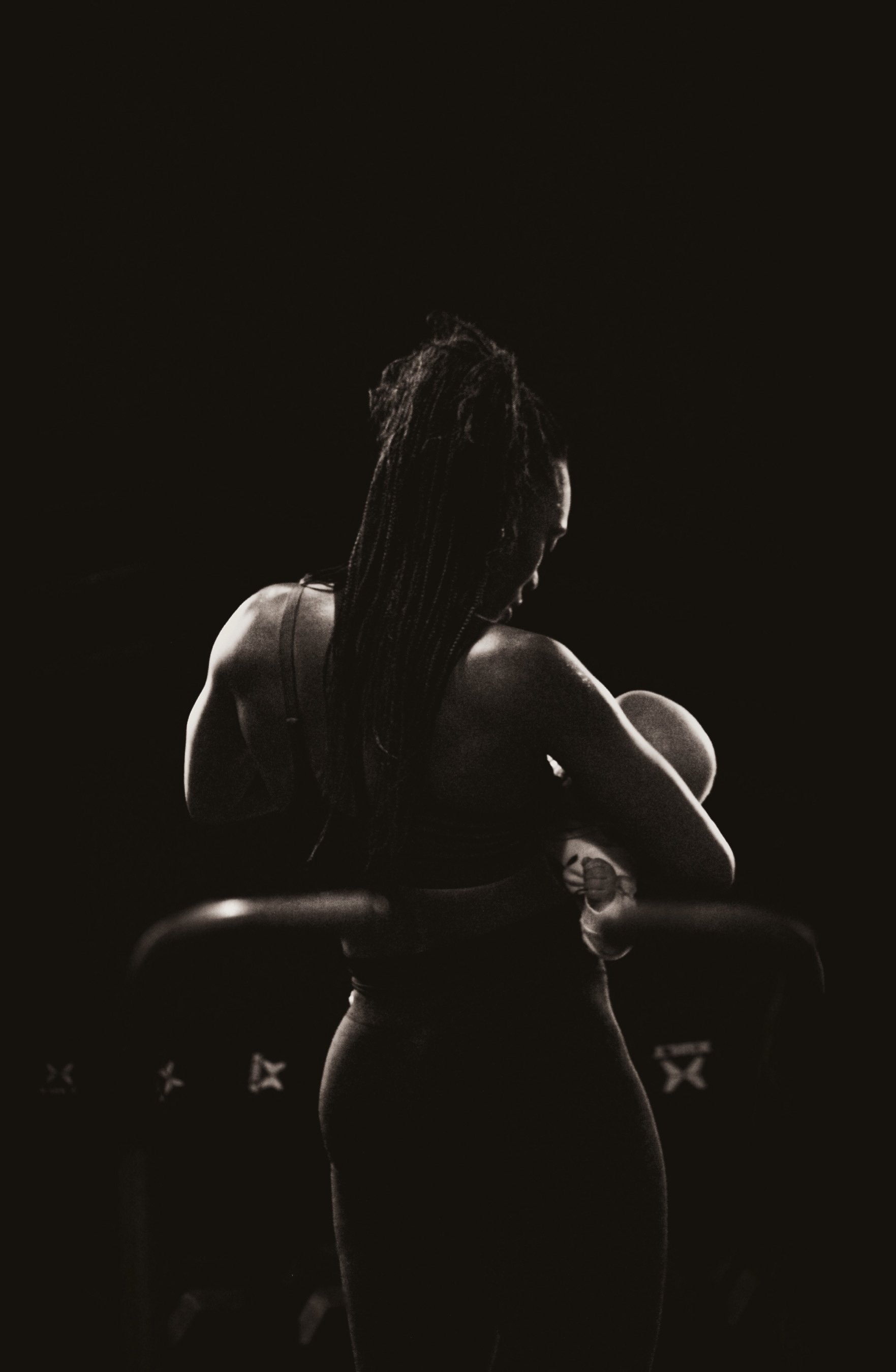 A black and white photo of a woman with dreadlocks hugging a baby, both facing to the right, in a dark setting with minimal lighting.