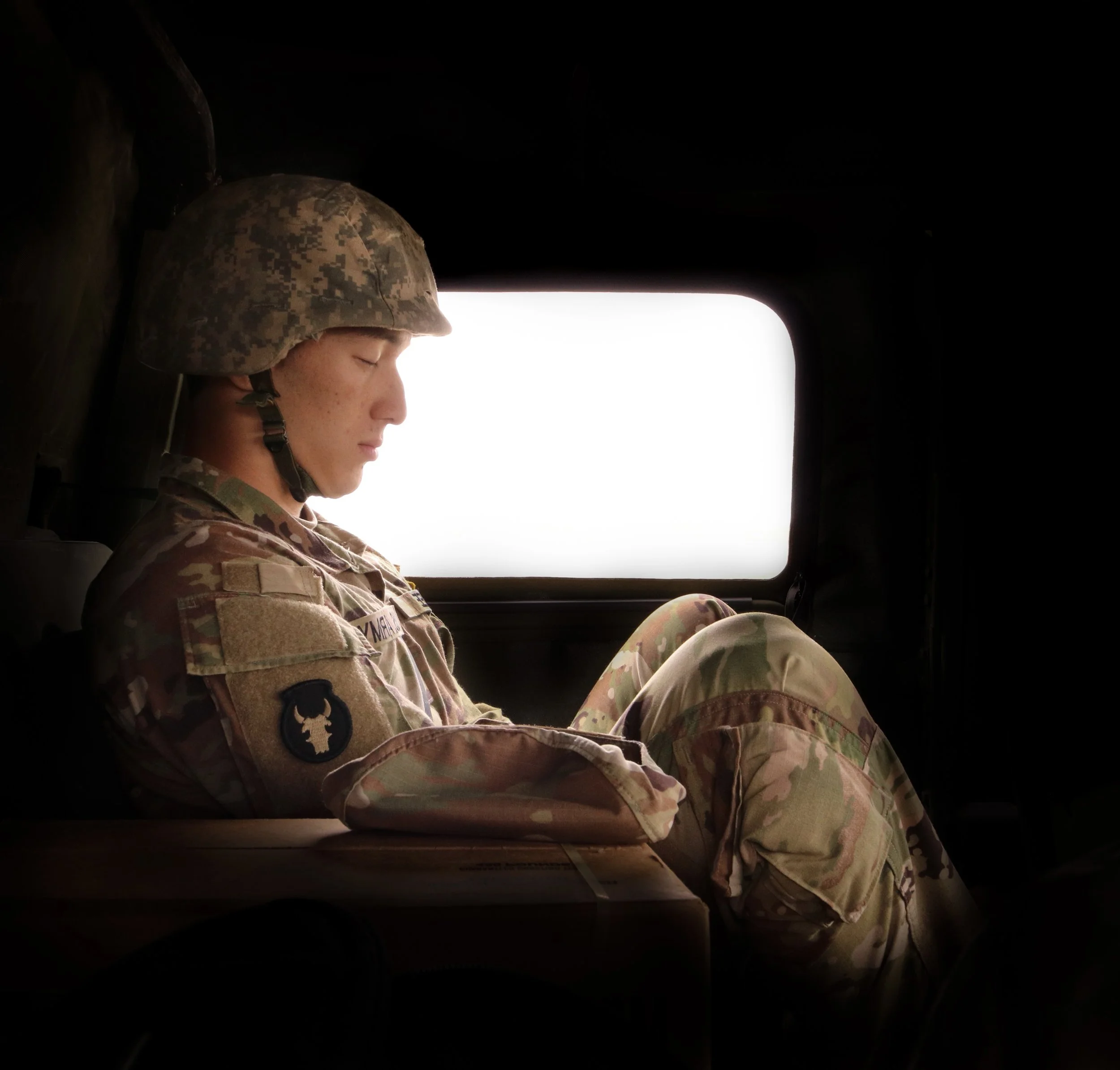 A soldier in camouflage uniform and helmet sitting with eyes closed inside a vehicle, with a bright white window behind.