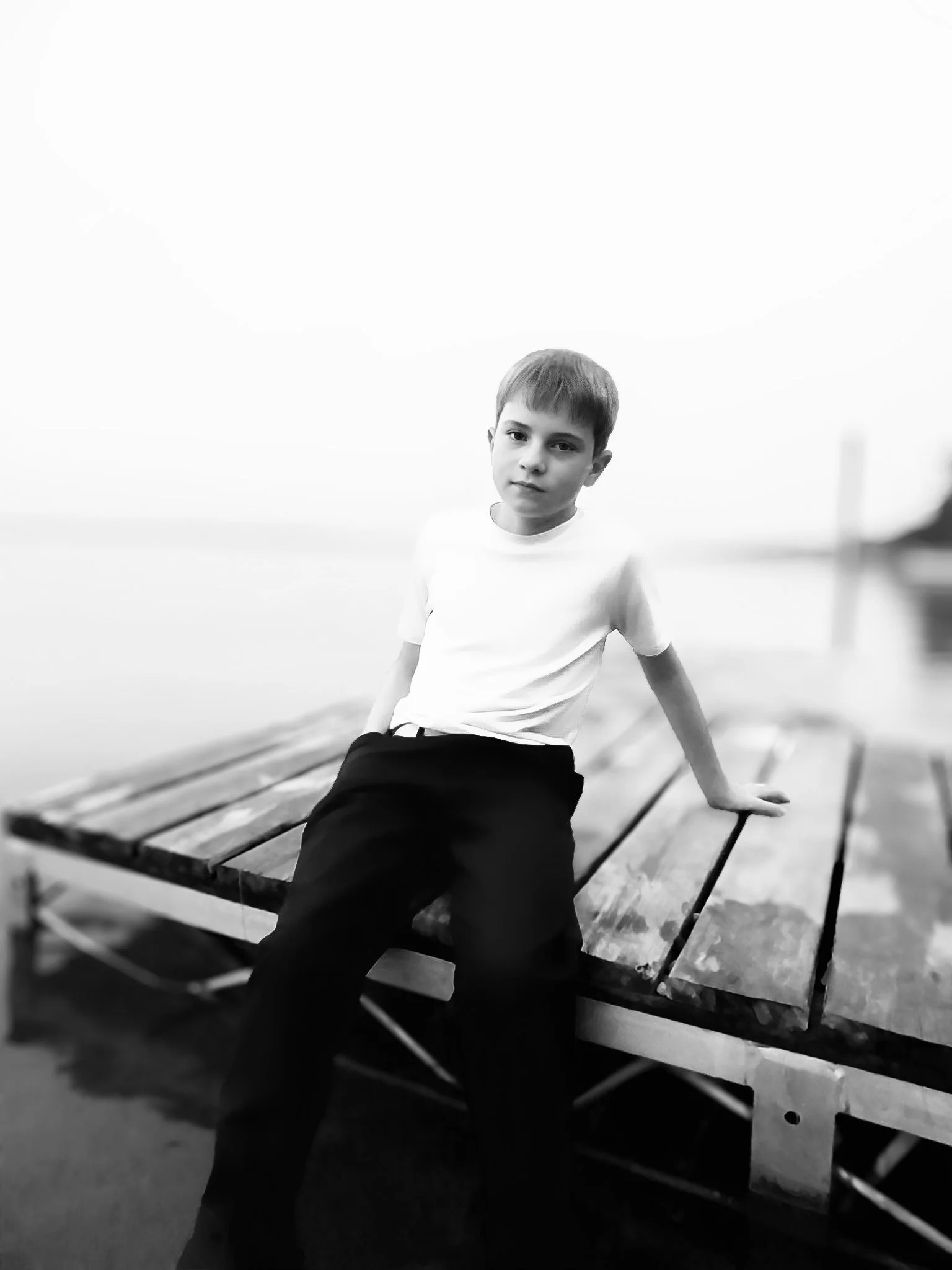 A young boy sitting on a wooden dock near the water with a blurred background, dressed in a white T-shirt and dark pants, looking at the camera.