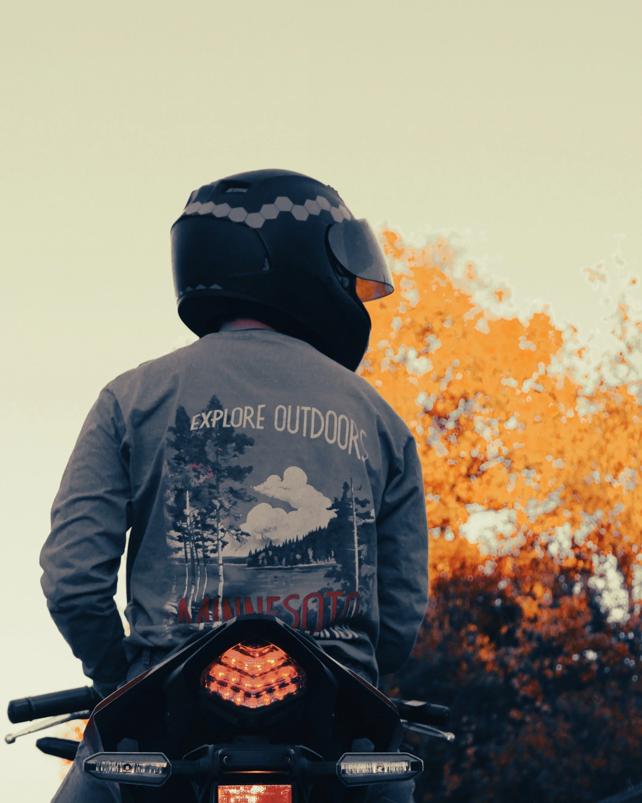 Person wearing a helmet and gray jacket on a motorcycle, with a forest fire in the background.