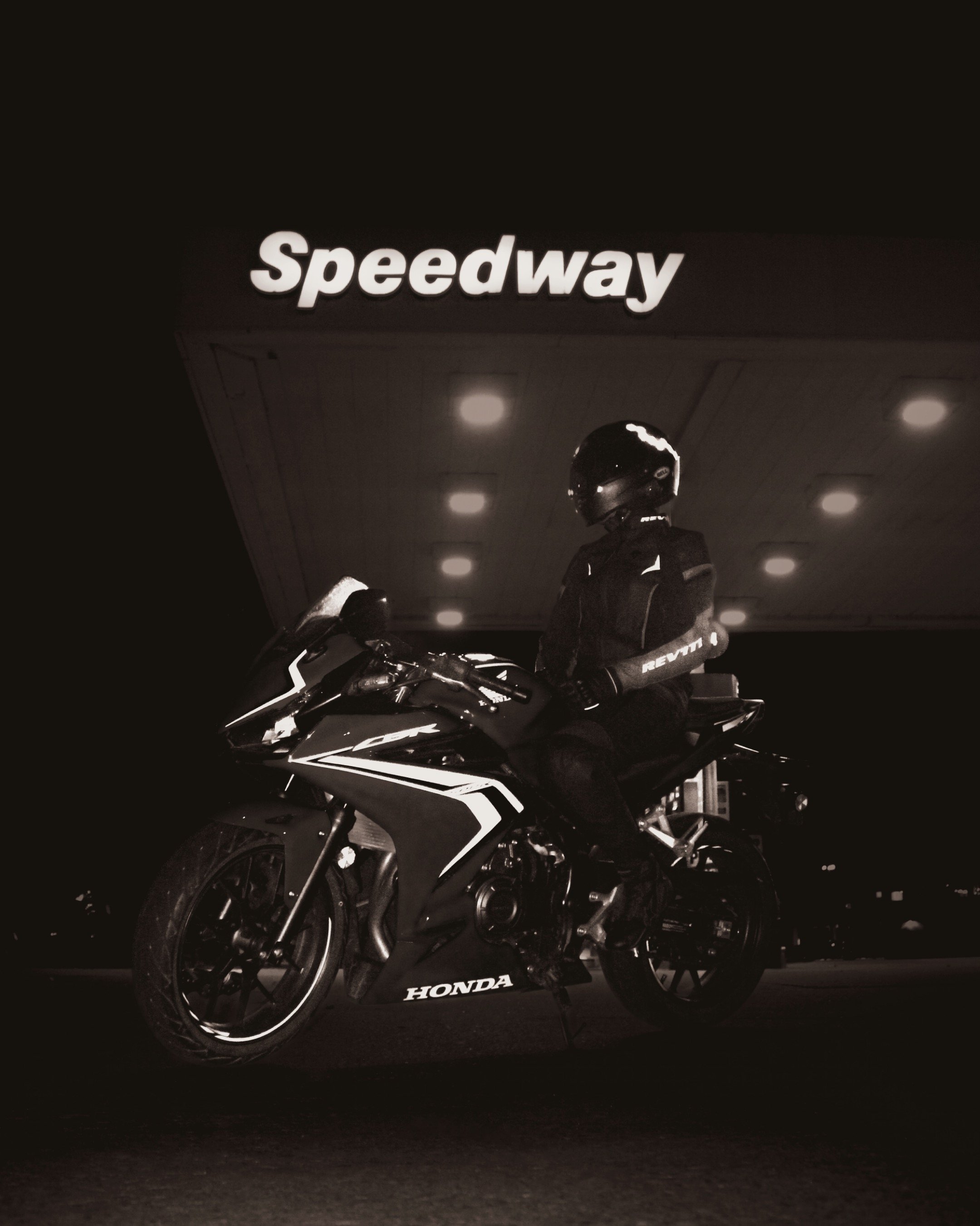 A motorcyclist wearing a helmet and riding gear sitting on a black Honda sport motorcycle at night under a Speedway sign.