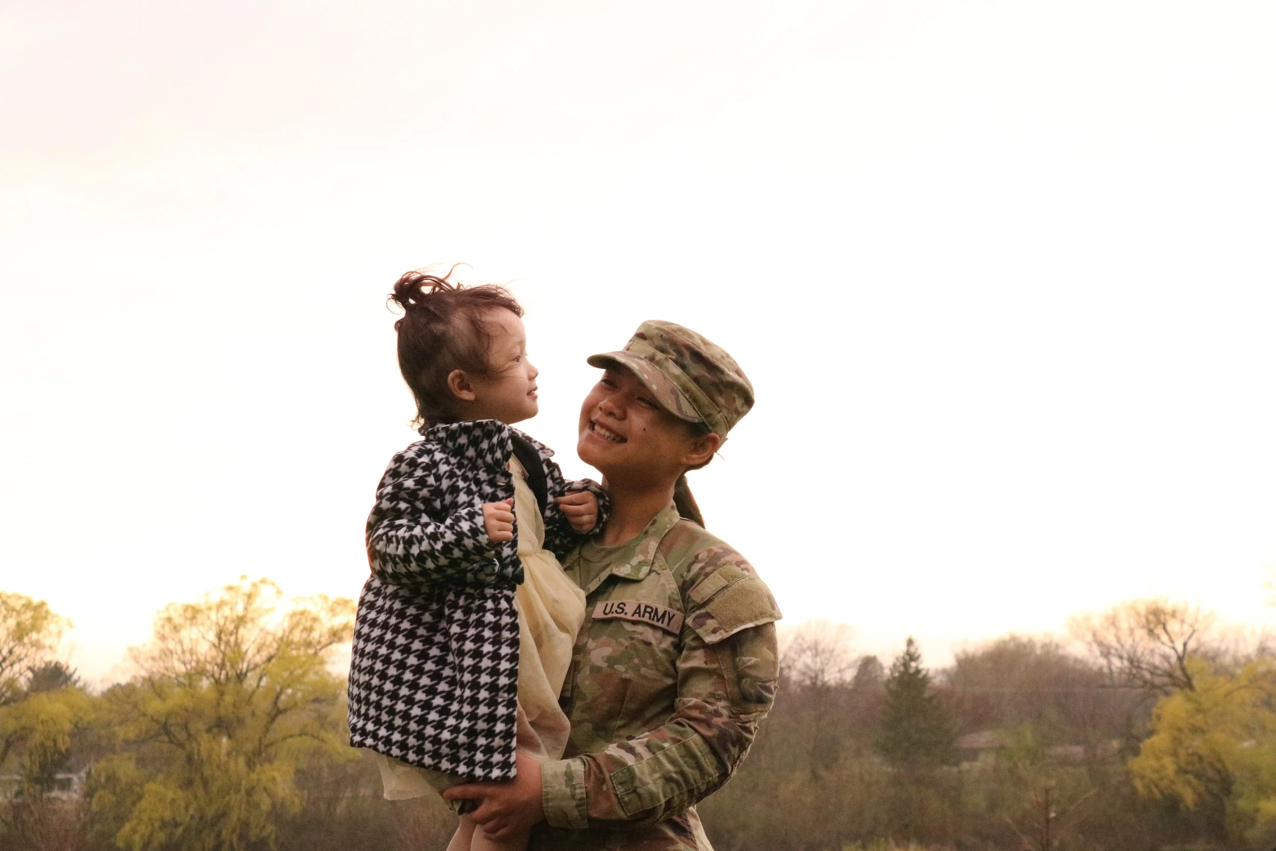 U.S. Army soldier holding a young girl in their arms outdoors with trees in the background.