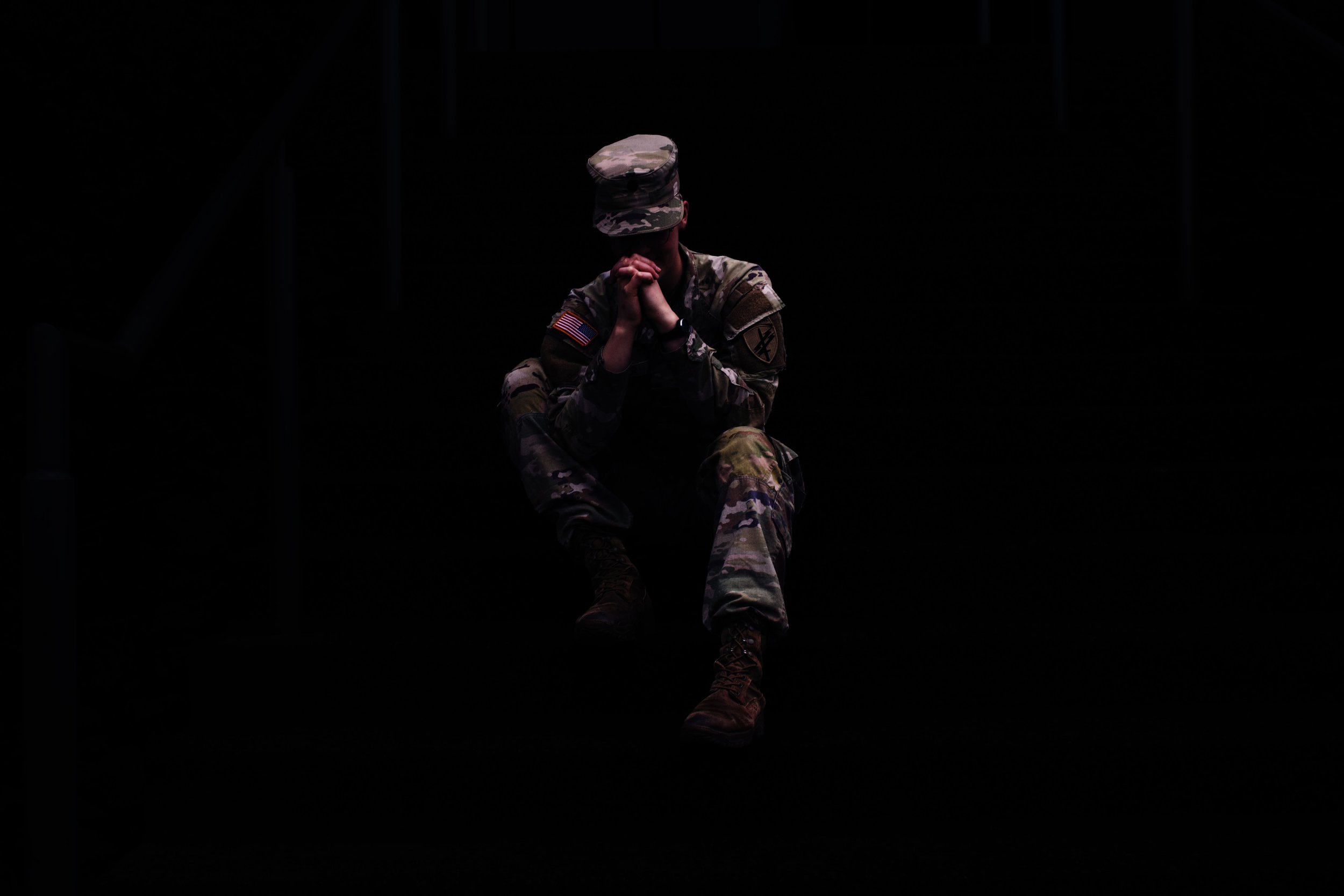 A soldier in camouflage uniform sitting with hands clasped and head bowed in prayer or contemplation.