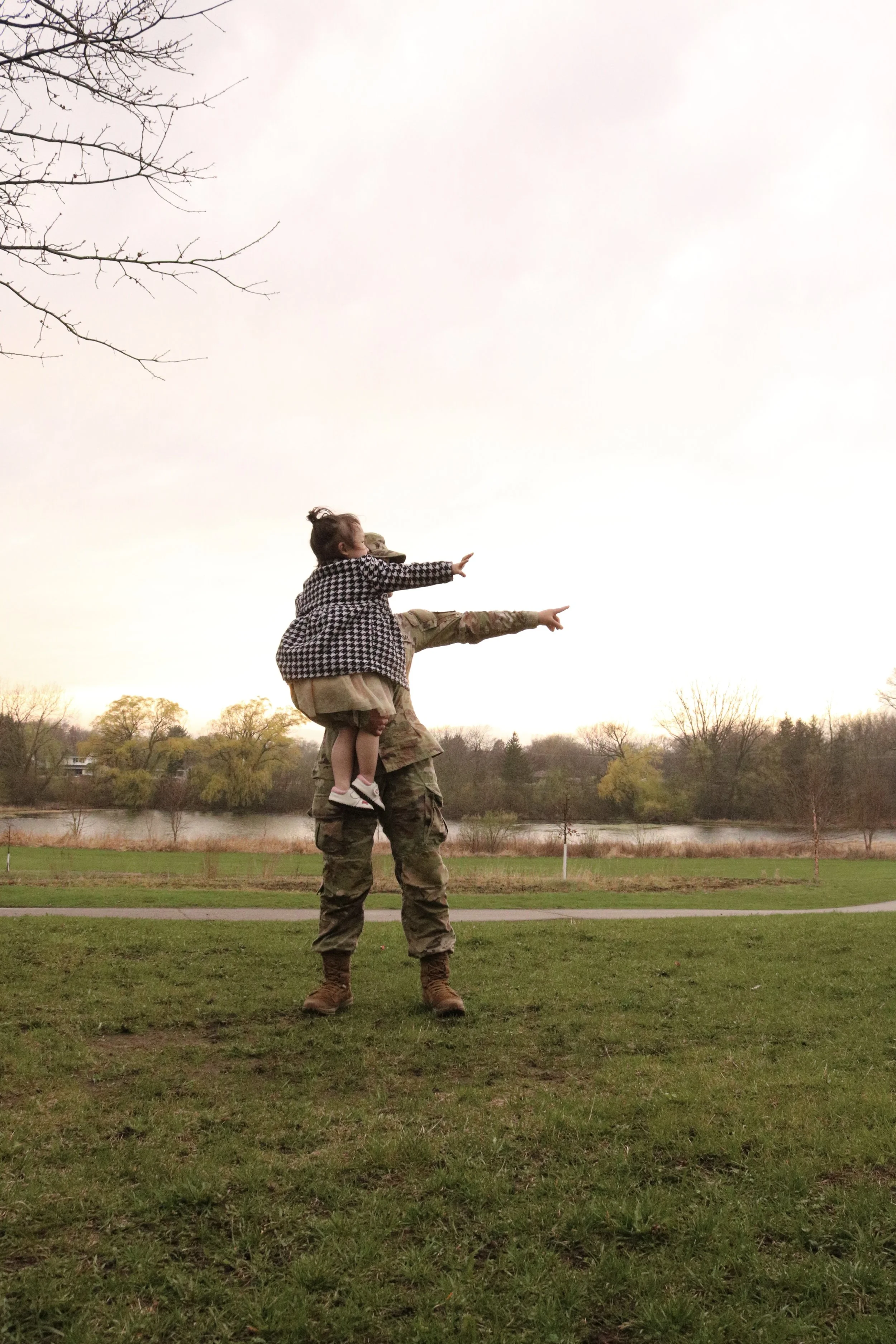 A female soldier standing on grass carrying a young girl on her shoulders, both pointing towards the sky in a park with a lake and trees in the background.